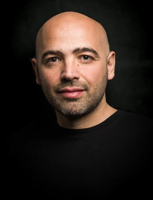 Head and shoulders portrait of photographer Costas Economou with a light beard wearing a black shirt, against a dark background.