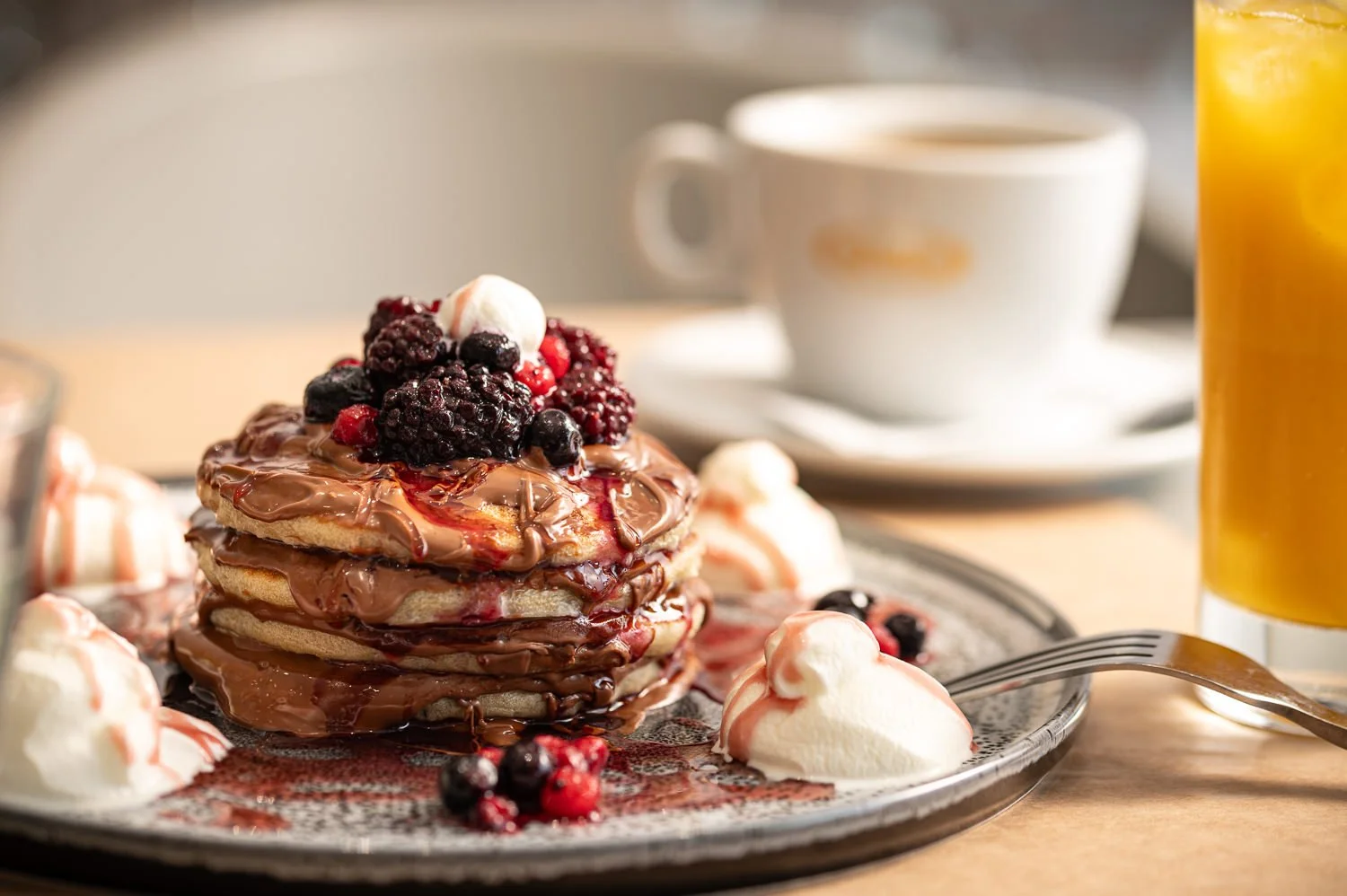 Stack of four pancakes topped with mixed berries and whipped cream, served with additional whipped cream, a cup of coffee, and a glass of orange juice on a wooden table. Hotels, Villas, Airbnb photography. Corfu, Greece.