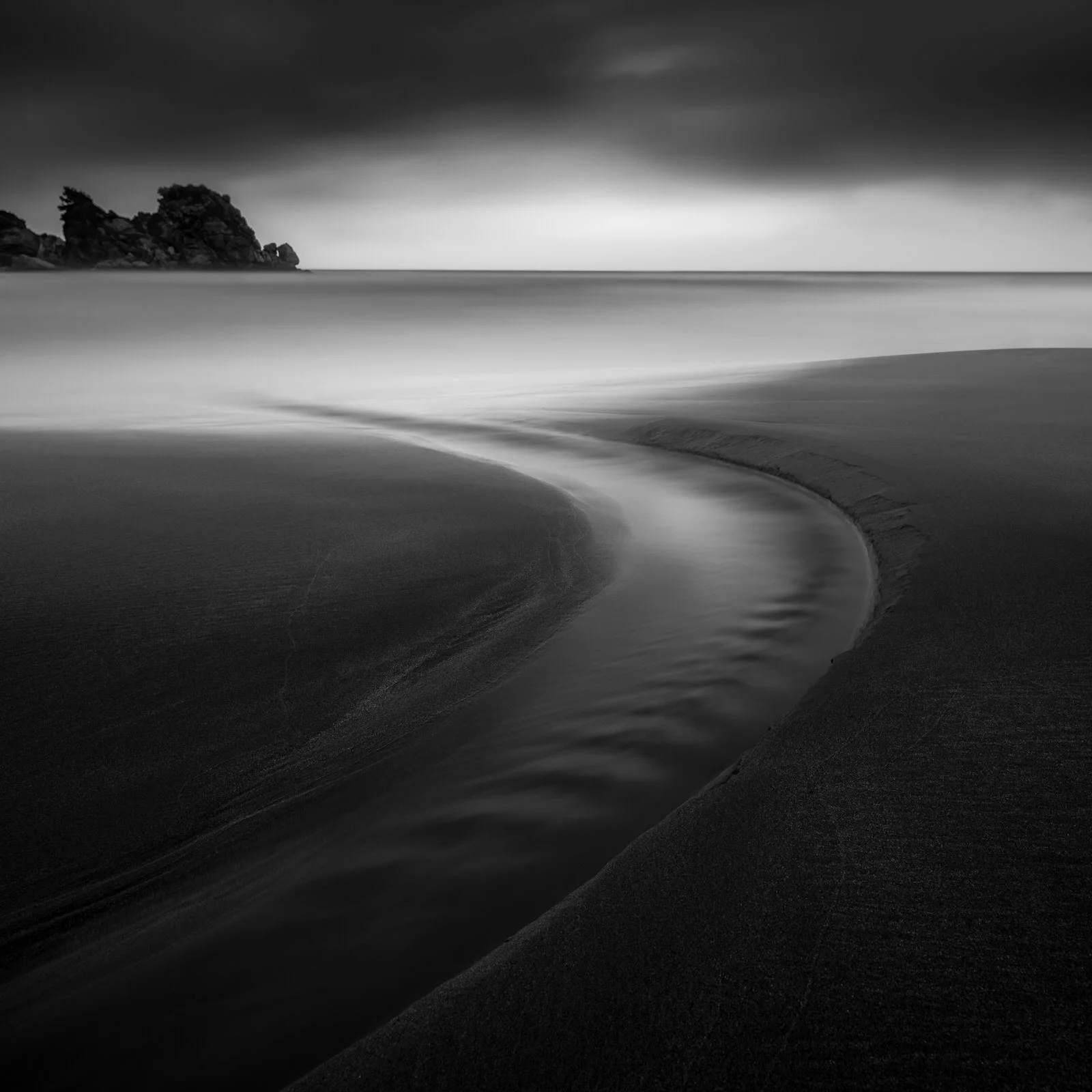 Black and white photo of a beach with a curved stream of water flowing into the ocean, with rocks in the distance under a cloudy sky.