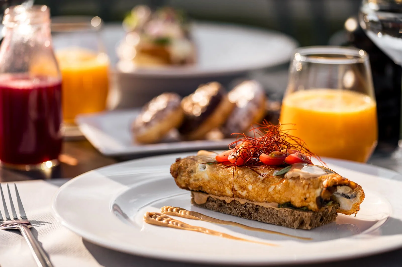 A breakfast plate with an omelet topped with chili threads and garnishes, In the background, there are glasses of orange juice, a bottle of juice, a plate of donuts, and a blurred outdoor setting. Hotels, Villas, Airbnb photography. Corfu, Greece.