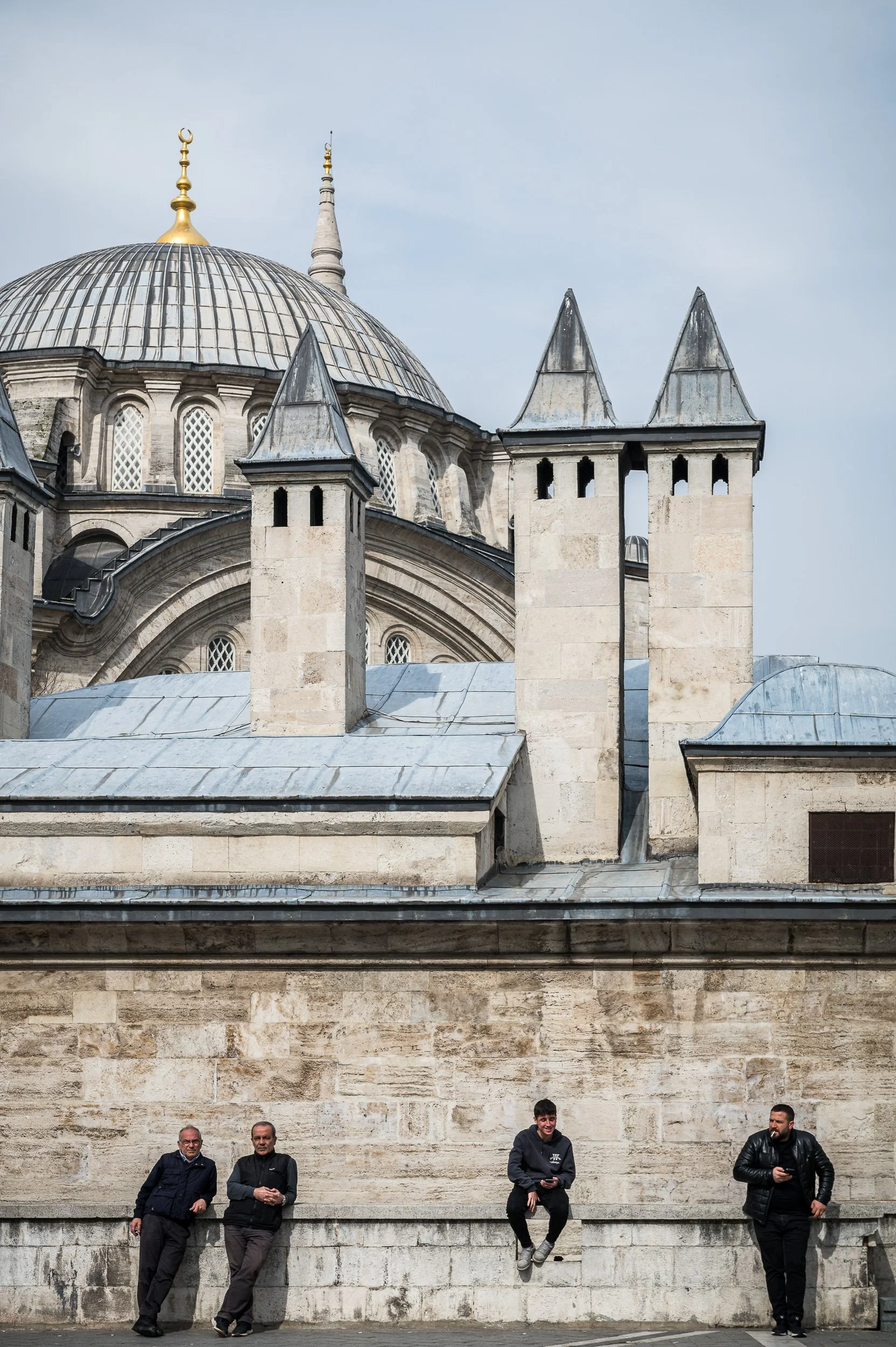 Four men standing and sitting in front of a historic stone building with domes and turrets, likely a mosque, under a partly cloudy sky.