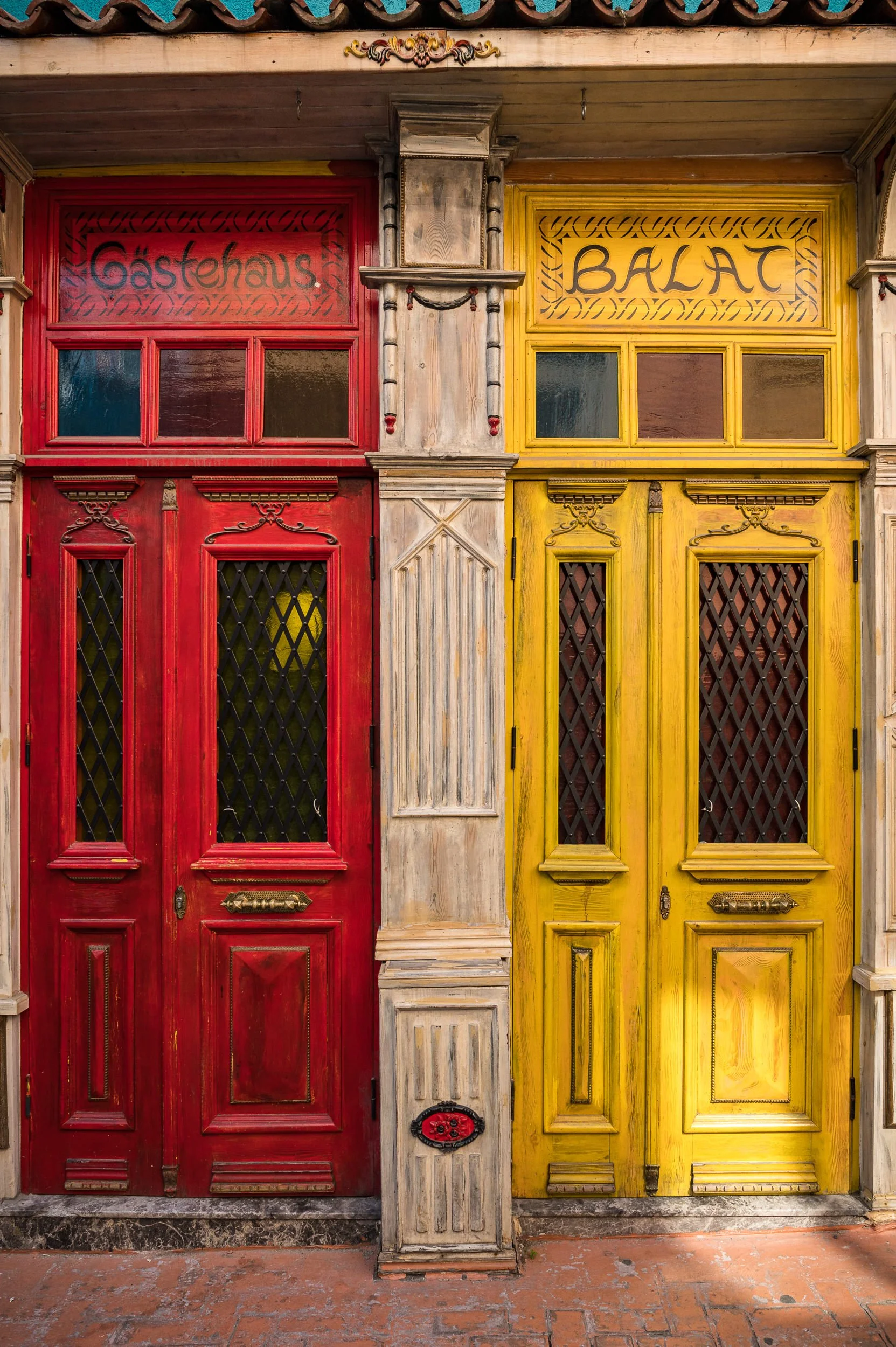 Bright red and yellow wooden doors with decorative trim and black lattice windows, labeled 'Gastehaus' and 'BALI' respectively, with a central column and vintage details.