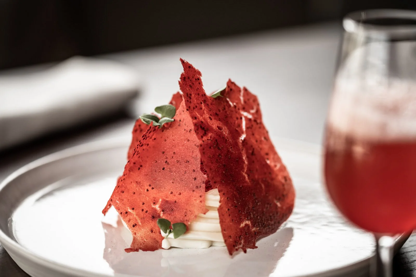 Elegant dessert with red spiral wafers, garnished with microgreens, served on a white plate with a blurred glass of pink-colored beverage in the background. Hotels, Villas, Airbnb photography. Corfu, Greece.