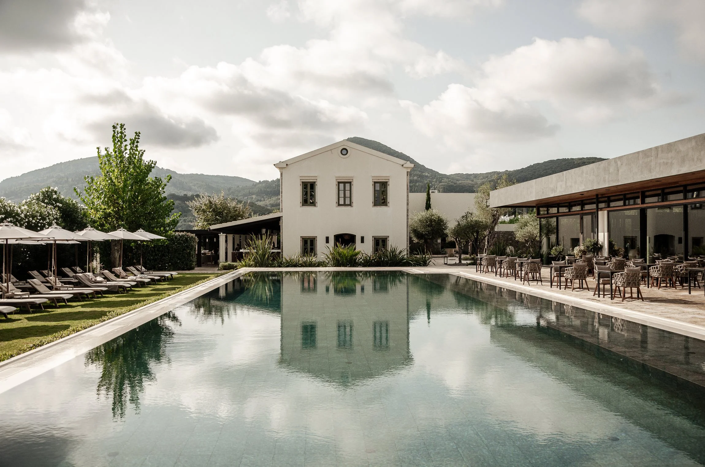 A luxury resort with a rectangular swimming pool, surrounded by lounge chairs and umbrellas, a white building in the background, and mountains beyond under a partly cloudy sky. Hotels, Villas, Airbnb photography. Corfu, Greece.