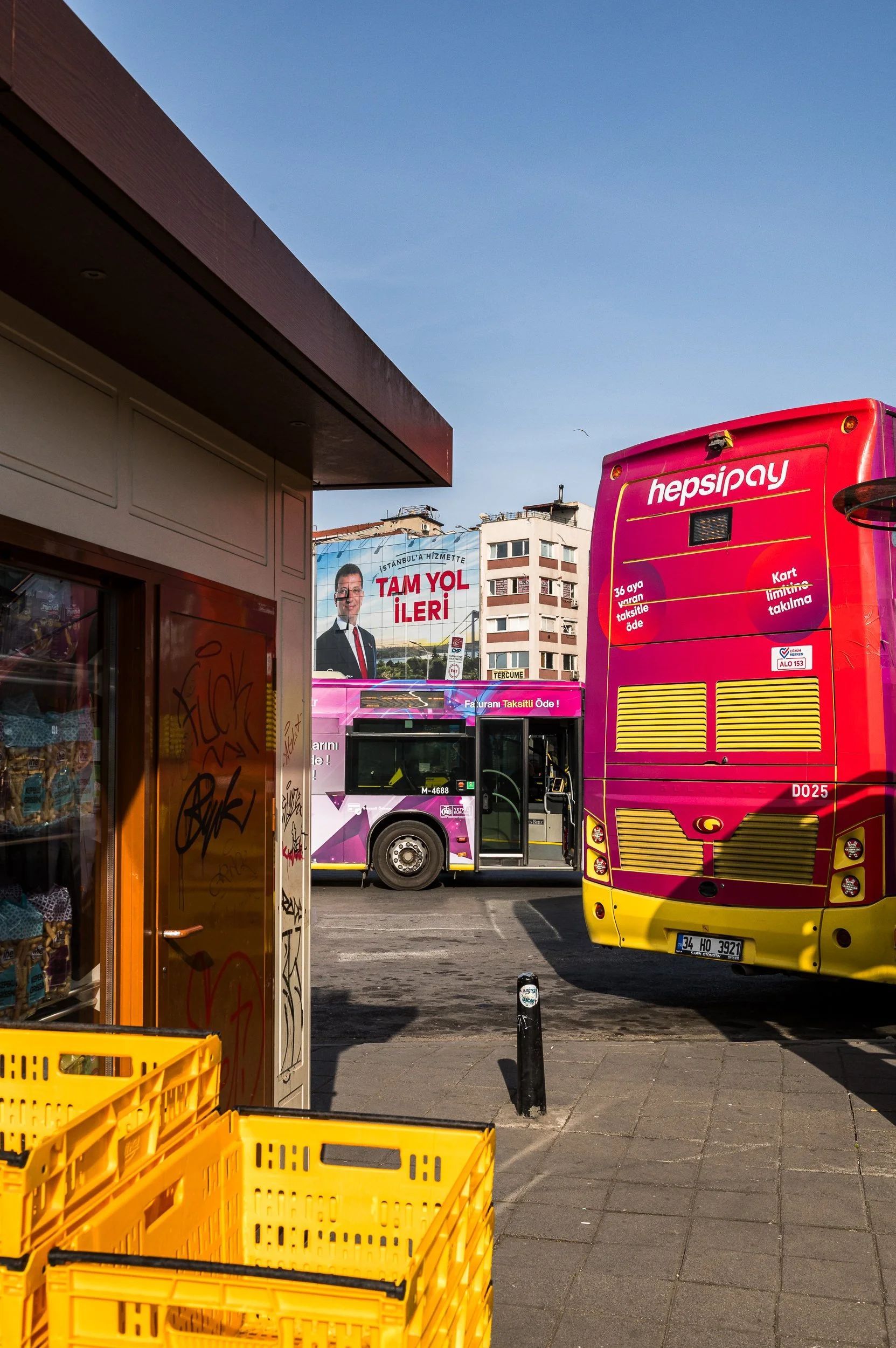 A street scene with pink and yellow buses, a small shop with yellow crates, and a billboard of a man in a suit with Turkish text in the background.