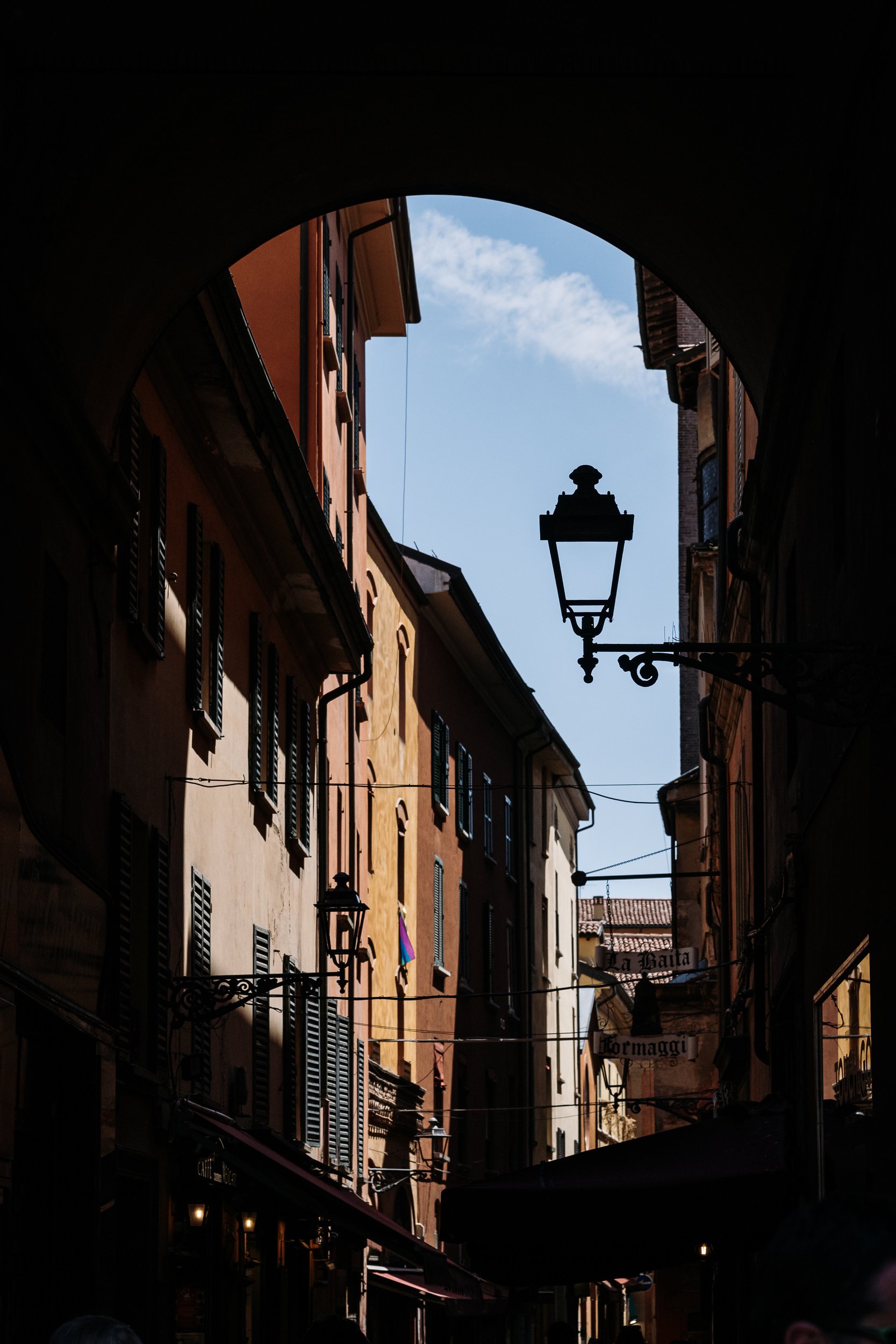 View of a narrow European street seen through an archway with old buildings, hanging signs, and street lamps, under a partly cloudy sky.