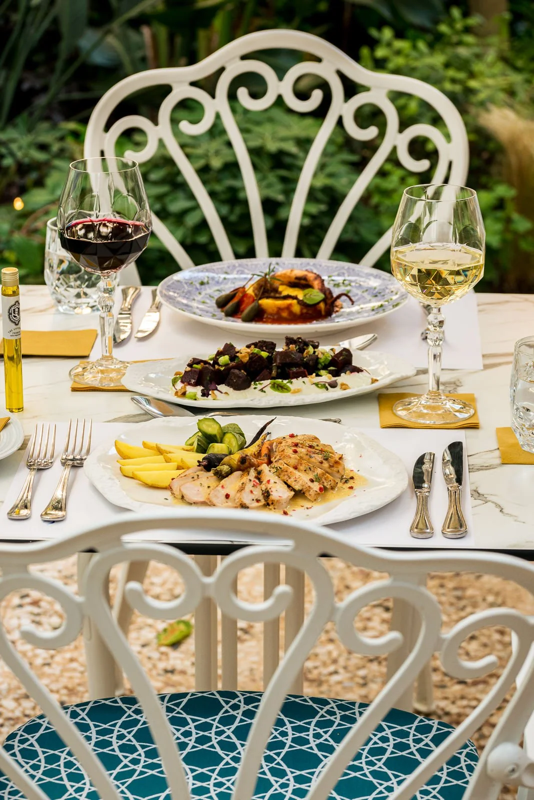 Elegant outdoor dining table set with white marble top, ornate white metal chairs, and dishes including sliced fish with lemon, grilled vegetables, and a beet salad, alongside glasses of red and white wine. Hotels, Villas, Airbnb photography. Corfu, 