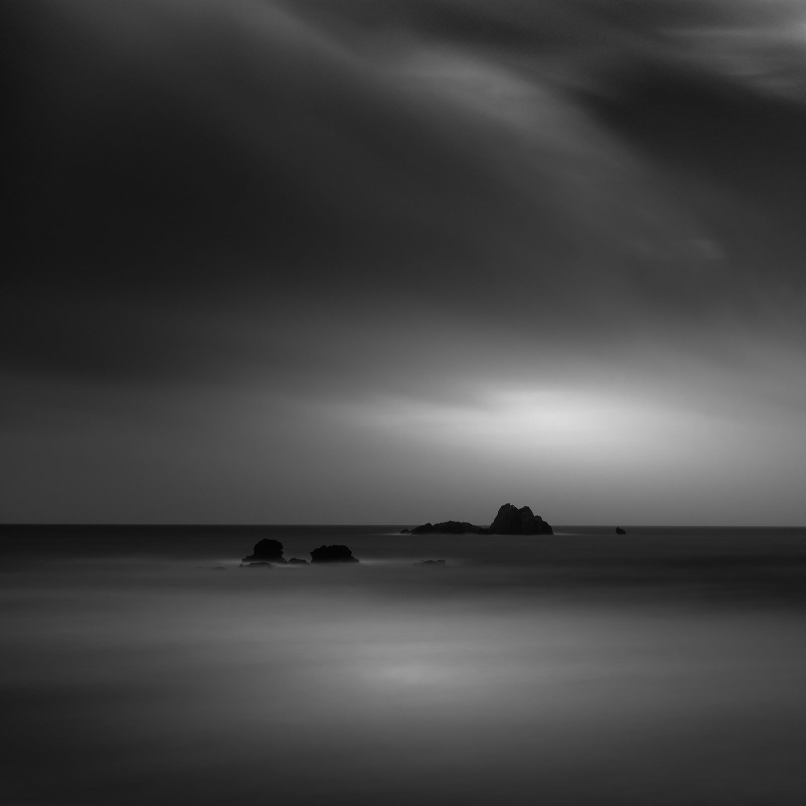 A black and white long exposure photograph of the ocean with rocks and a cloudy sky.