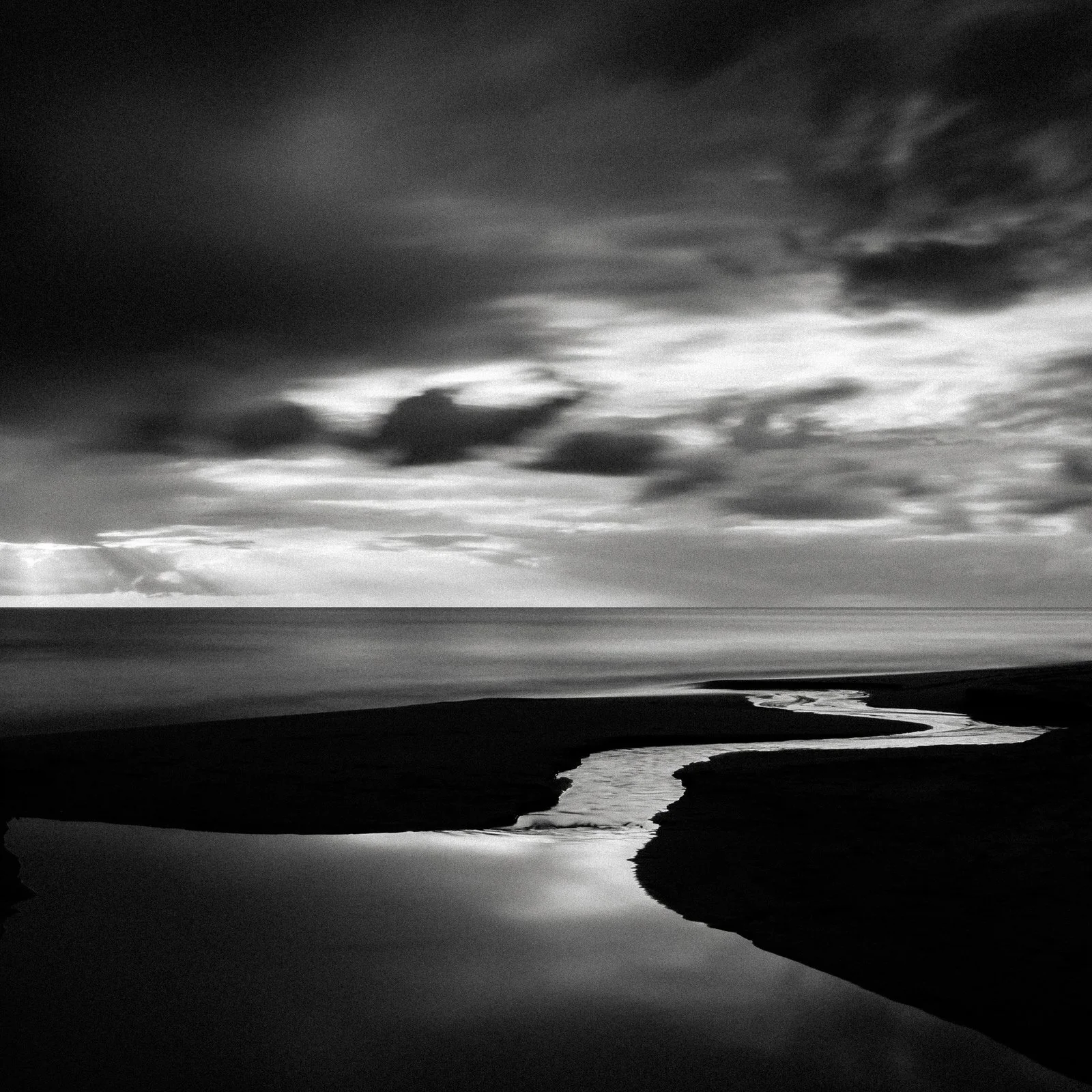Black and white photograph of a coastal scene with a winding stream flowing into the ocean under a cloudy sky.