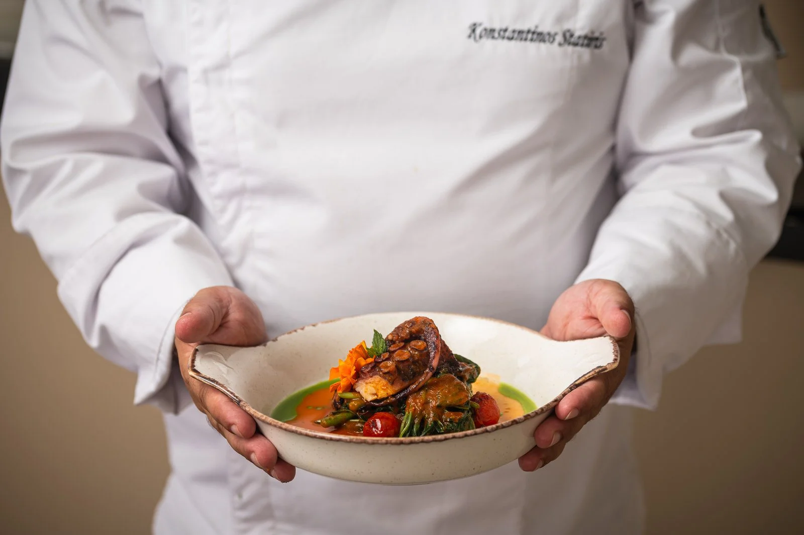 Person in a white chef's coat holding a ceramic bowl with a plated gourmet dish, including meat, vegetables, and colorful sauce. Hotels, Villas, Airbnb photography. Corfu, Greece.