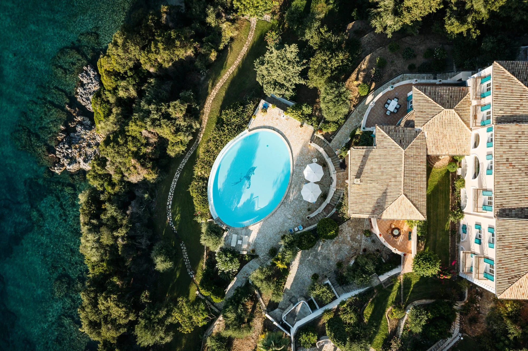 Aerial view of a large estate with a kidney-shaped swimming pool, landscaped yard, and multi-story house with terracotta tile roof, situated near a body of water with rocky shoreline and trees. Hotels, Villas, Airbnb photography. Corfu, Greece.