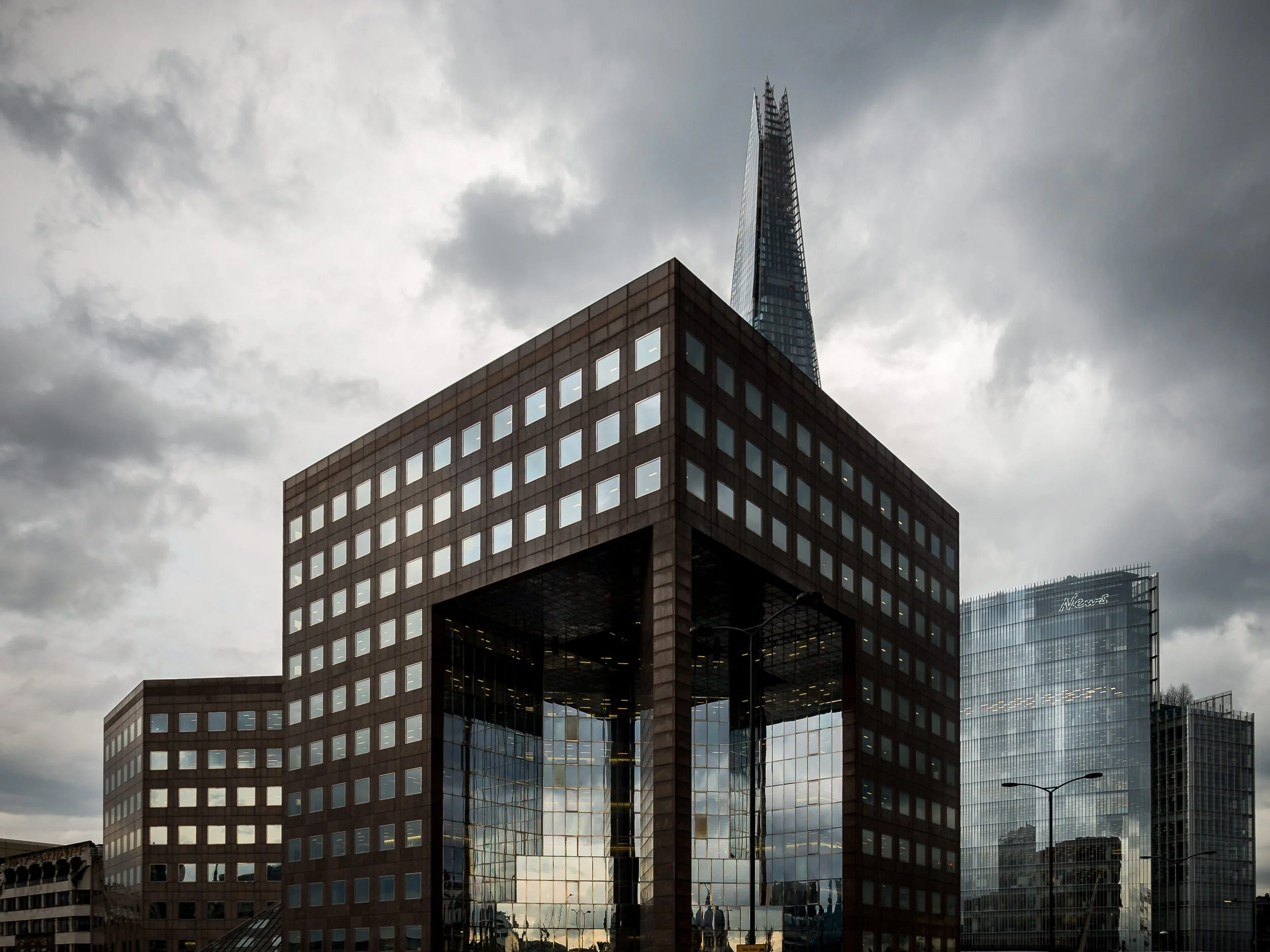 Modern cityscape with a large, brown office building featuring a glass-covered opening, and reflective windows, with the skyline including the Shard and other glass skyscrapers, under a cloudy sky.