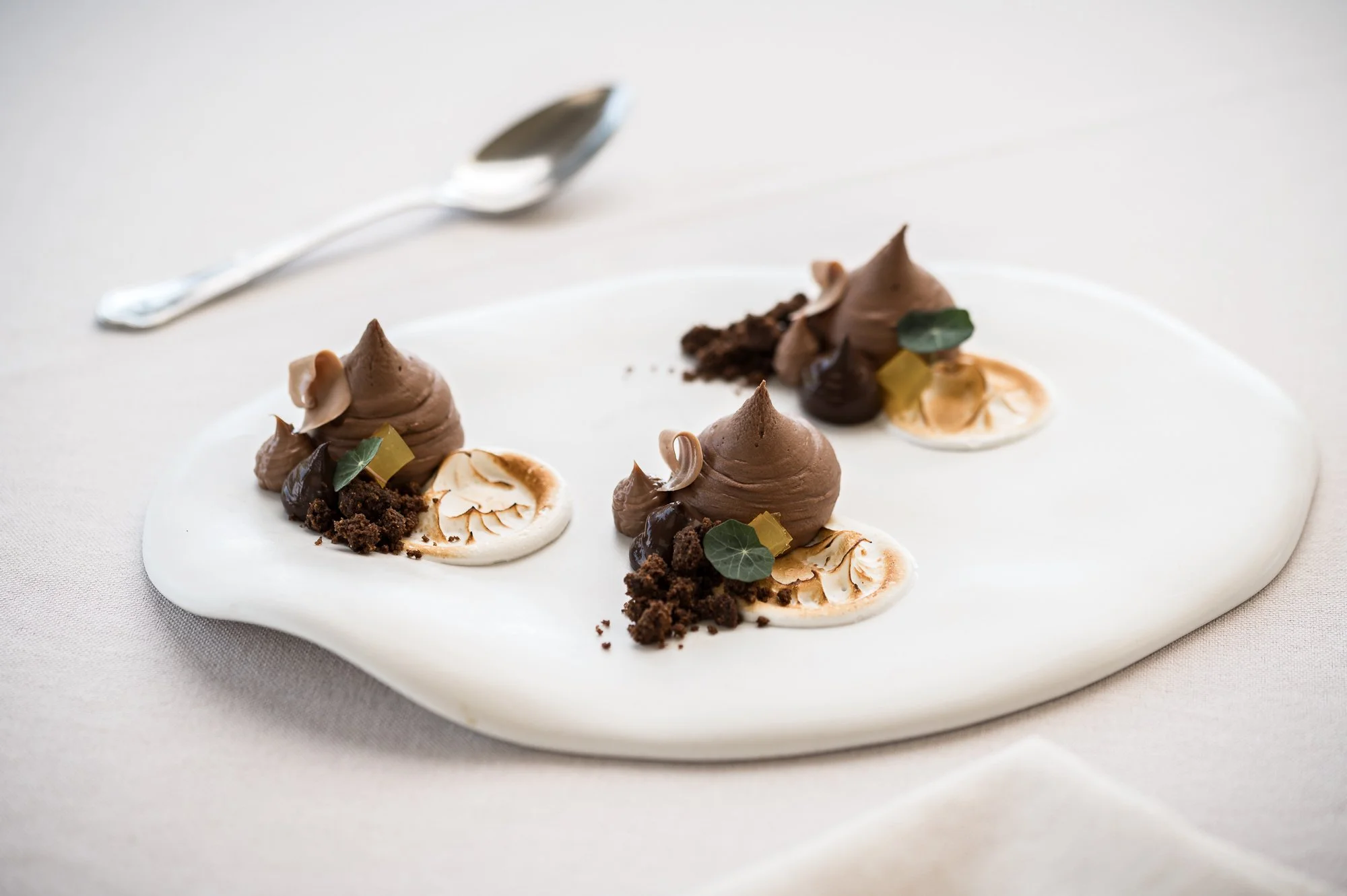 Three chocolate mousse desserts garnished with edible leaves, crumbs, and cream on a white irregular-shaped plate with a silver spoon in the background. Hotels, Villas, Airbnb photography. Corfu, Greece.