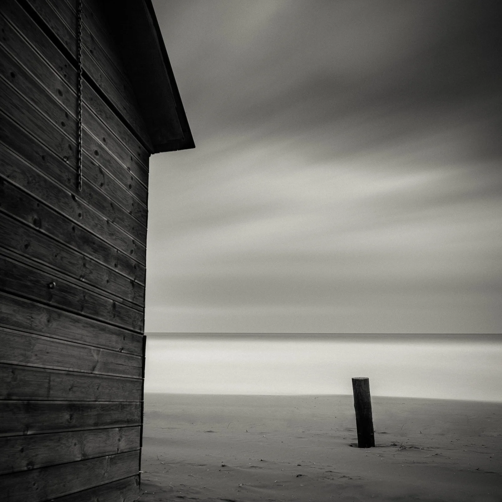 A black and white photo showing a wooden beach house on the left, a single leaning post on the sand, and a calm ocean in the background under a cloudy sky.