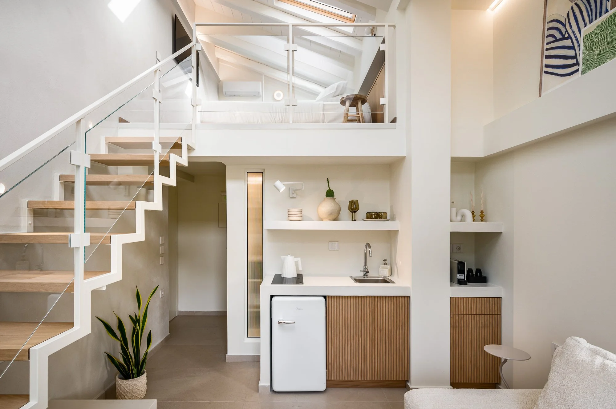 Interior of a modern, compact apartment with a loft bed above a small kitchen area, featuring white walls, wooden accents, and decorative plants. Hotels, Villas, Airbnb photography. Paxos, Greece.