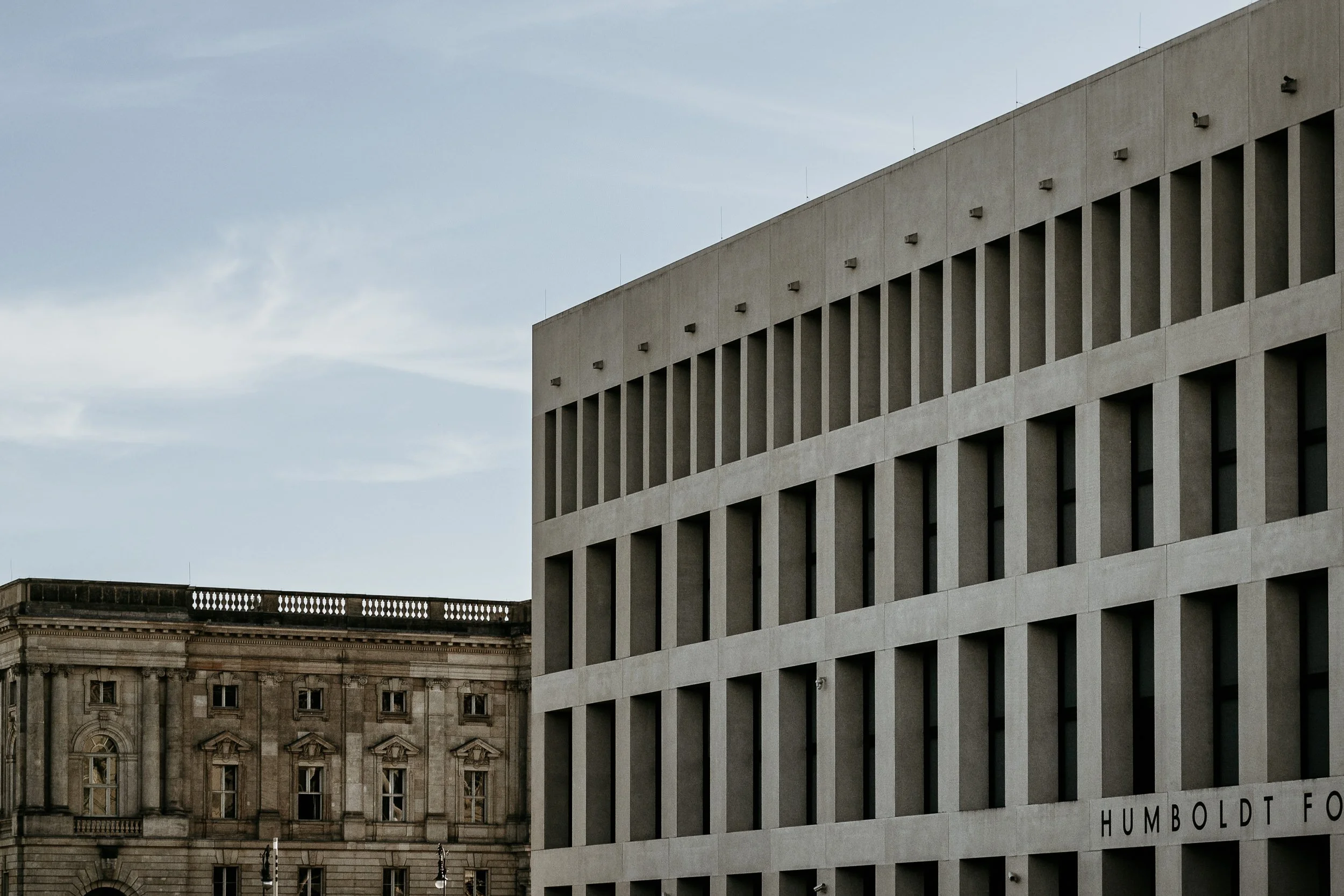 Modern beige multi-story building with evenly spaced windows on the facade, next to a historic stone building with ornate window frames, against a partly cloudy sky.
