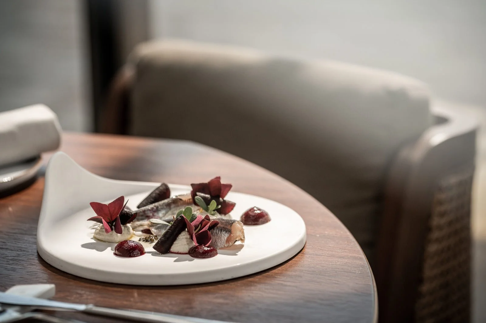 A plated gourmet dish with fish fillet garnished with red edible flowers, sauce, and green herbs on a white ceramic plate, on a wooden table with chairs and a napkin nearby. Hotels, Villas, Airbnb photography. Corfu, Greece.