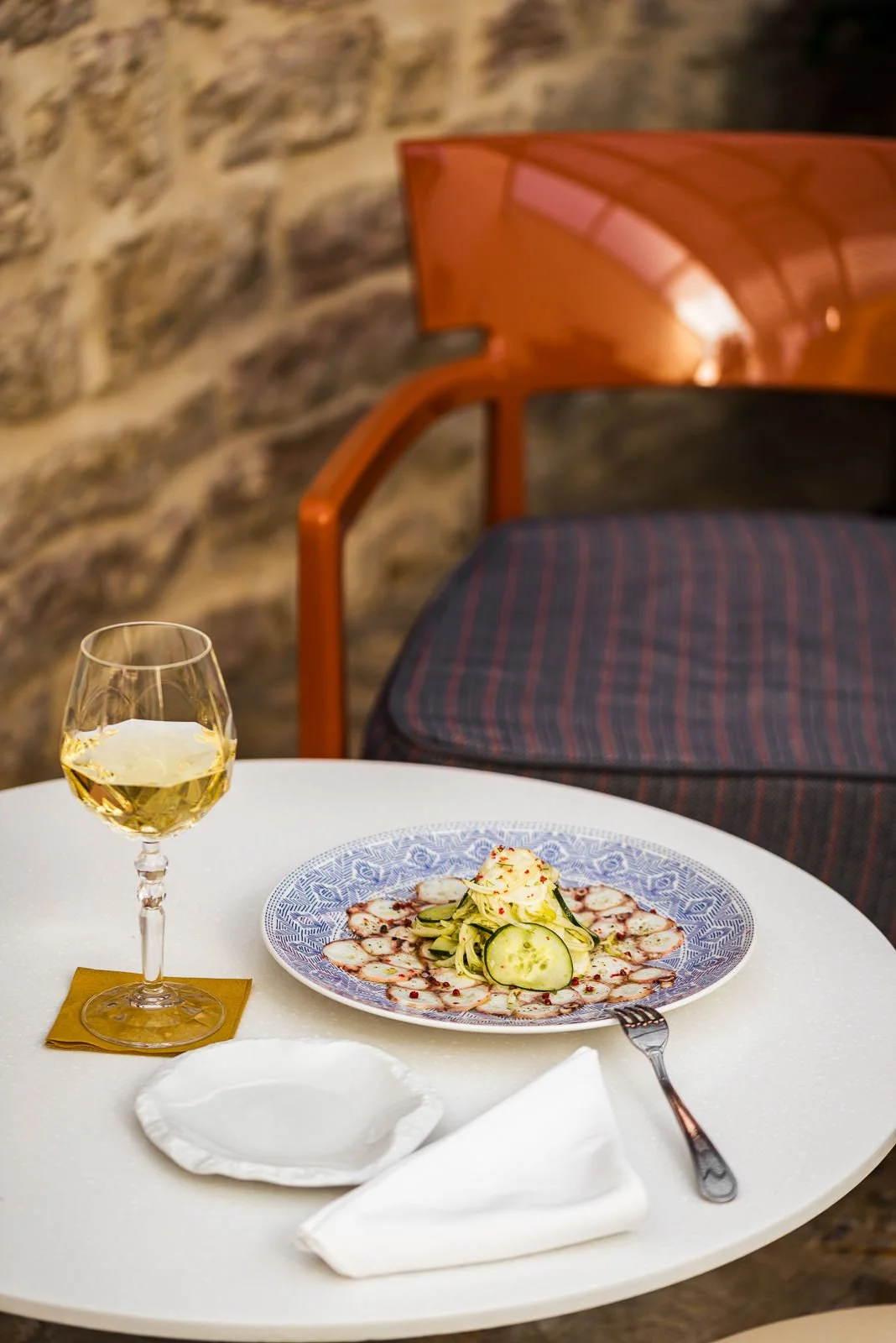 A table set with a glass of white wine, a plate of sliced cucumbers and radishes garnished with red pepper flakes. In the background, a wooden chair with a cushioned seat and a stone wall. Hotels, Villas, Airbnb photography. Corfu, Greece.