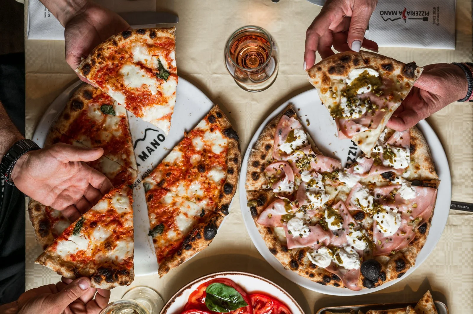 Two pizzas being served at a restaurant table, one with cheese and basil, the other with ham, cheese, and herbs, along with a glass of rosé wine and a bowl of sliced tomatoes. Hotels, Villas, Airbnb photography. Corfu, Greece.
