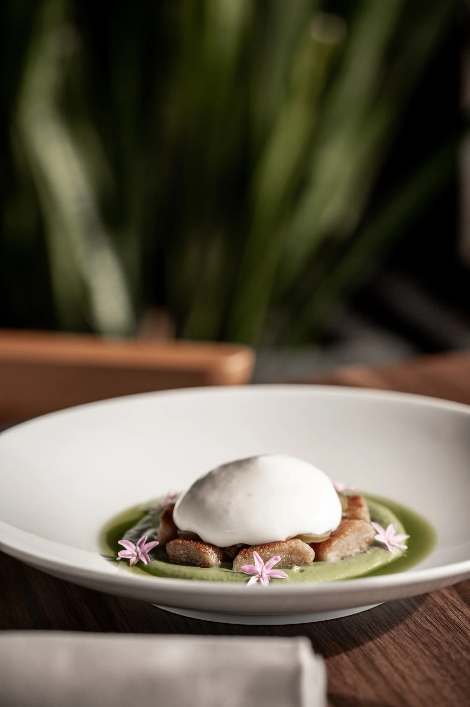 A plated dish with a poached egg on top of sliced cooked vegetables and green sauce, garnished with pink flowers on a white plate. Hotels, Villas, Airbnb photography. Corfu, Greece.