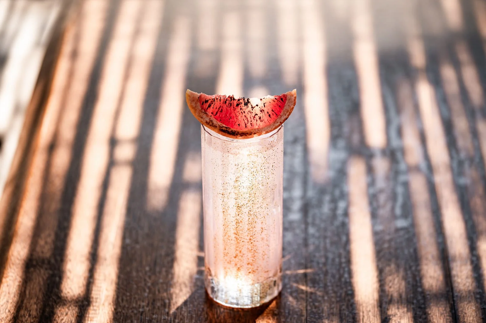 A tall, pink-colored cocktail with a slice of watermelon on top, sitting on a wooden surface with striped shadows.Hotels, Villas, Airbnb photography. Corfu, Greece.