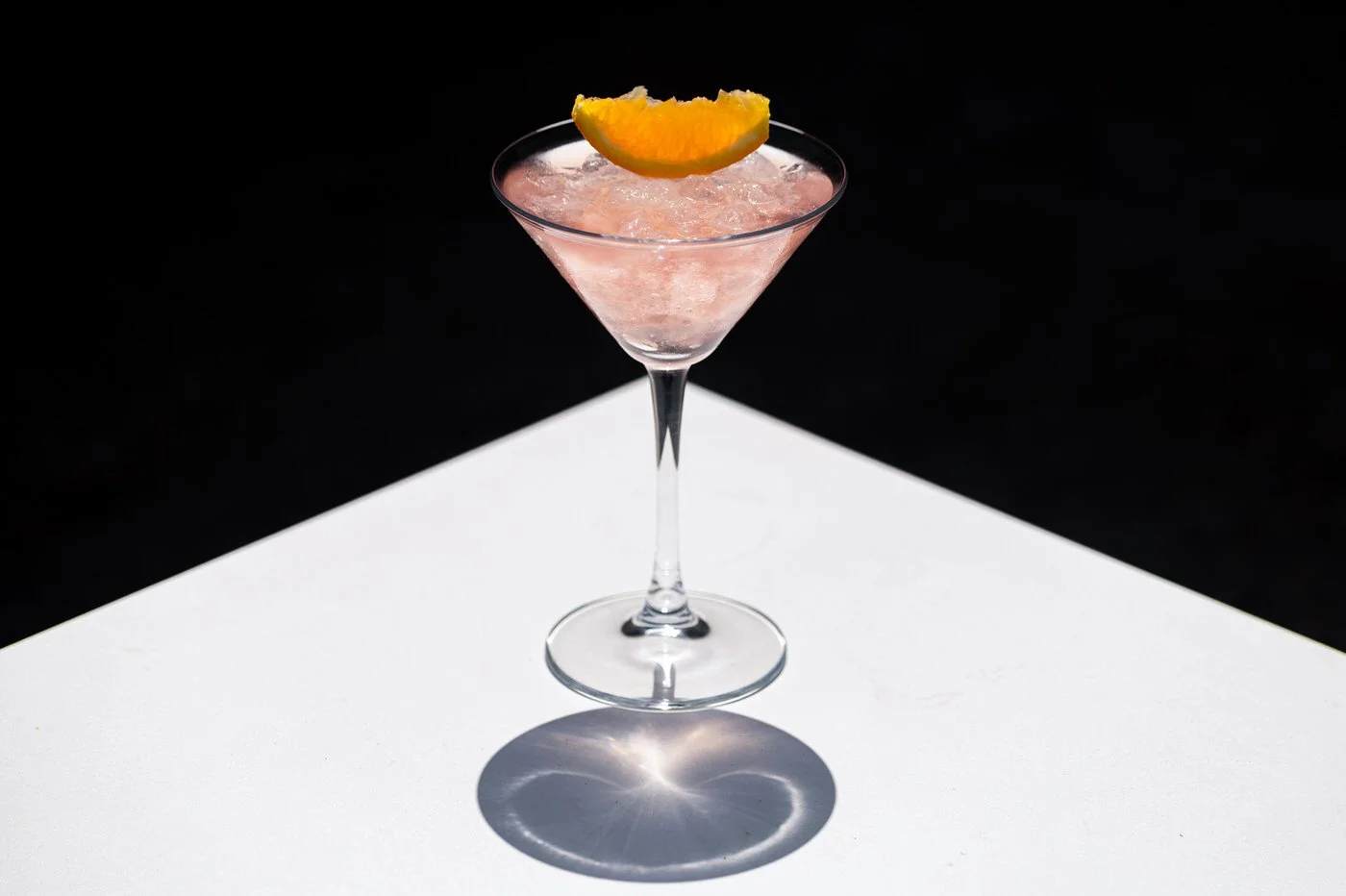Pink cocktail in a martini glass with an orange wedge garnish on top, against a black background with a white surface reflecting the glass. Hotels, Villas, Airbnb photography. Corfu, Greece.