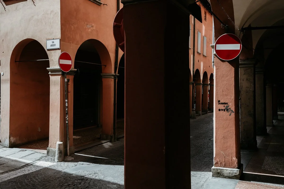 Street scene with orange buildings, archways, and two red no-entry traffic signs attached to columns, in an alleyway with cobblestone pavement.