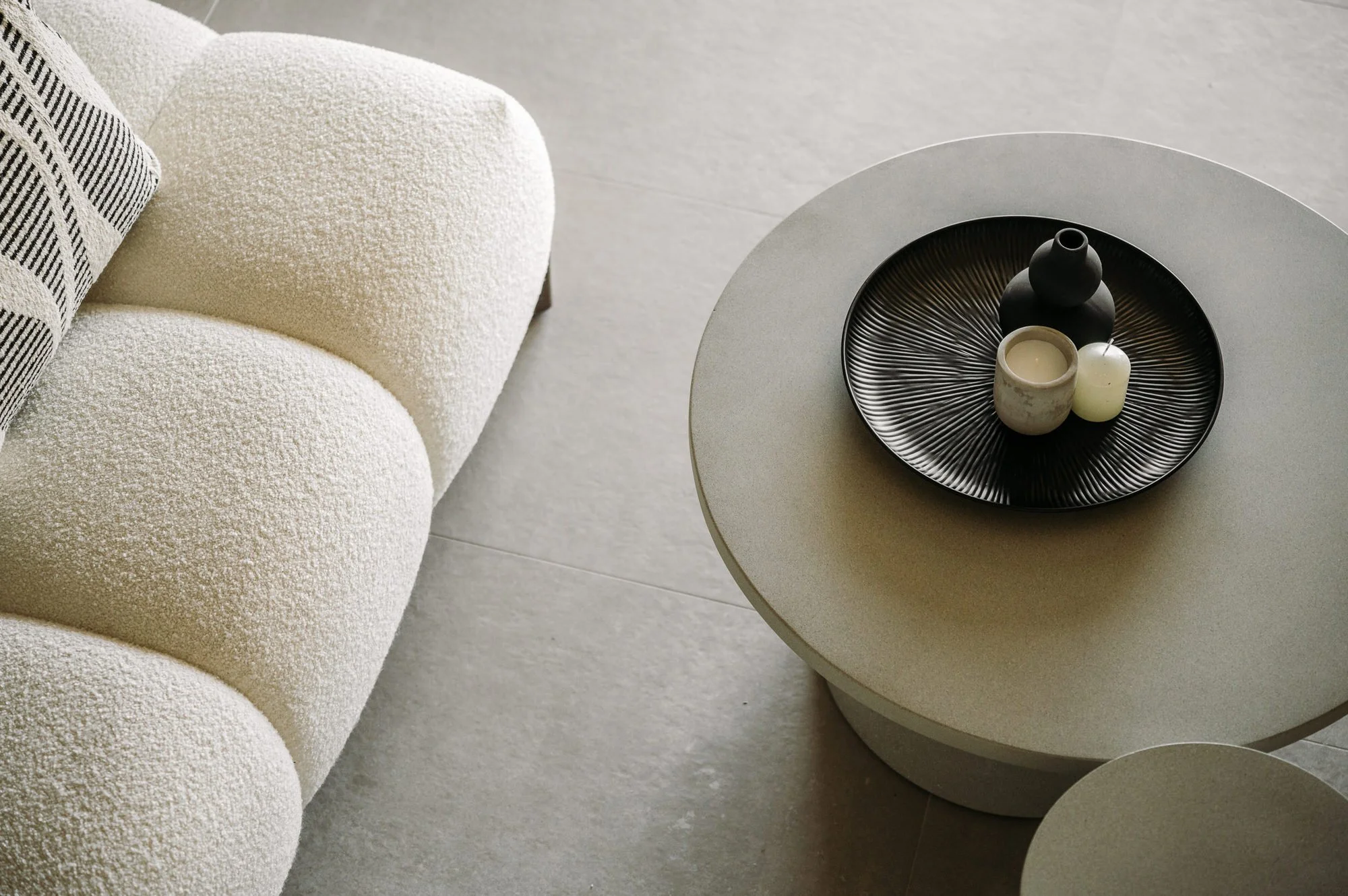 Close-up of a textured cream-colored sofa with a black and white striped pillow and a round beige coffee table with a black decorative tray holding three candles and a black vase. Hotels, Villas, Airbnb photography. Corfu, Greece.
