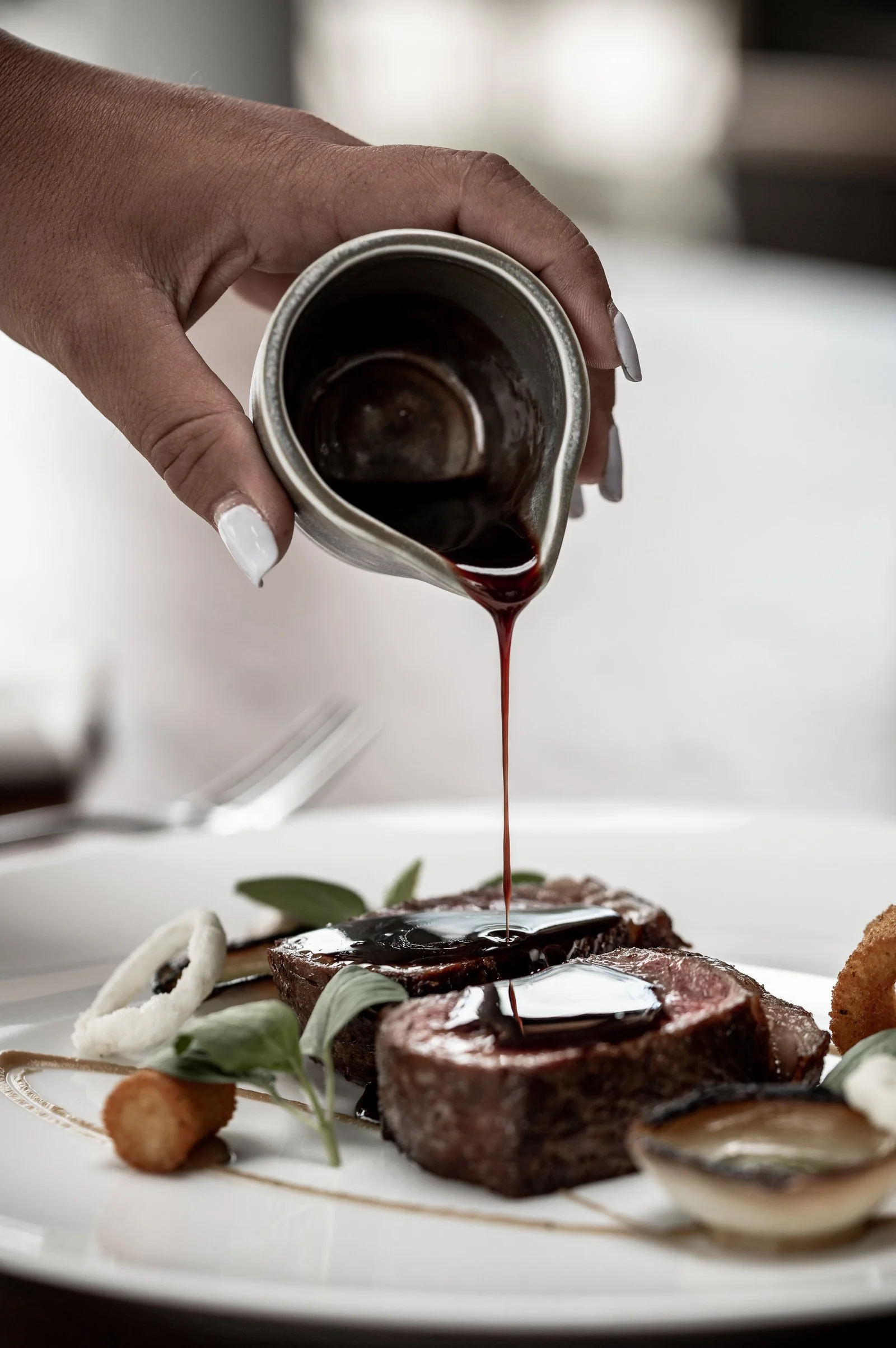 A person pouring dark sauce over two cooked steaks on a plate, garnished with herbs, onions, and fried items. Hotels, Villas, Airbnb photography. Corfu, Greece.
