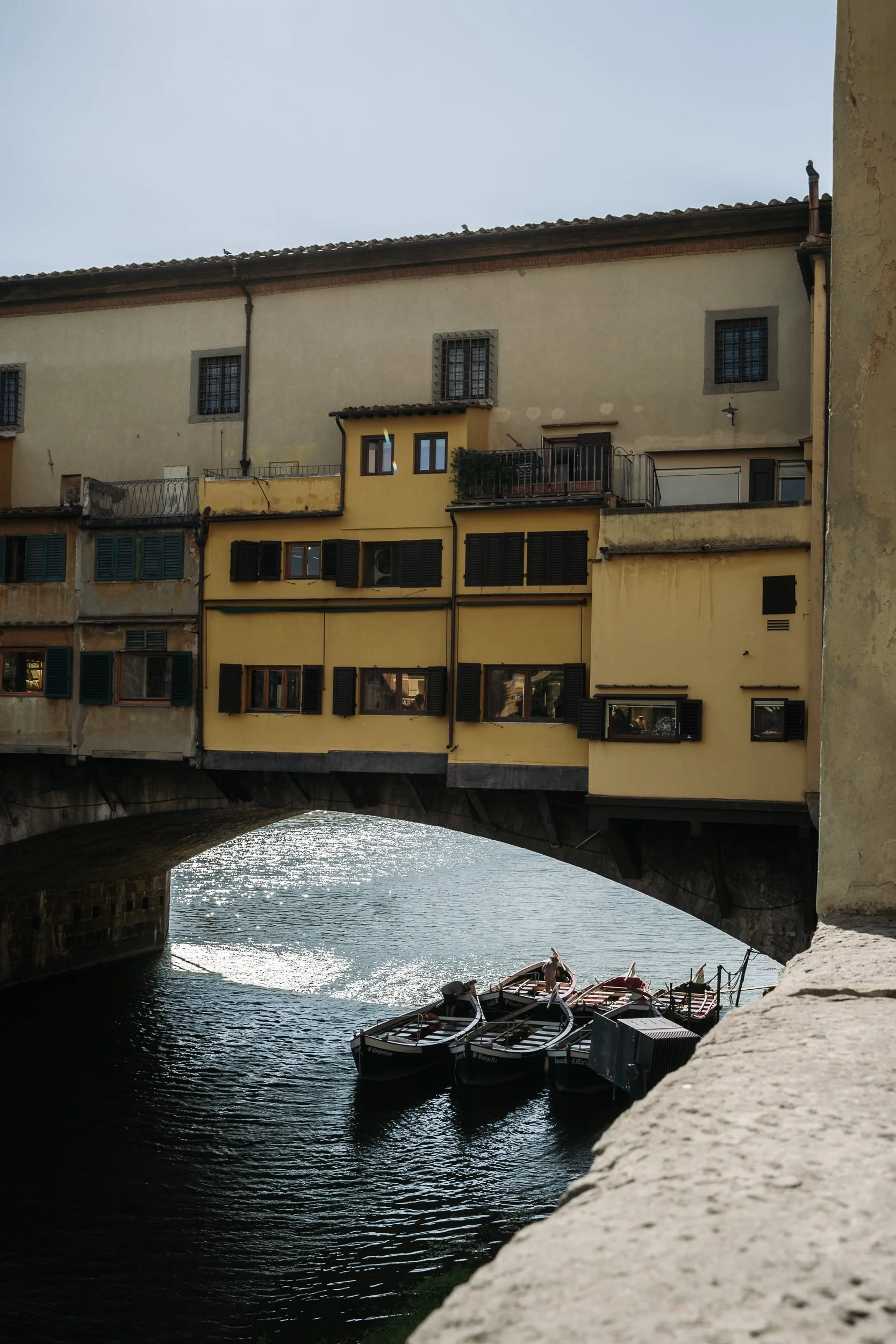 Colorful buildings with yellow and beige facades and green shutters over a small canal with boats, under a bridge.