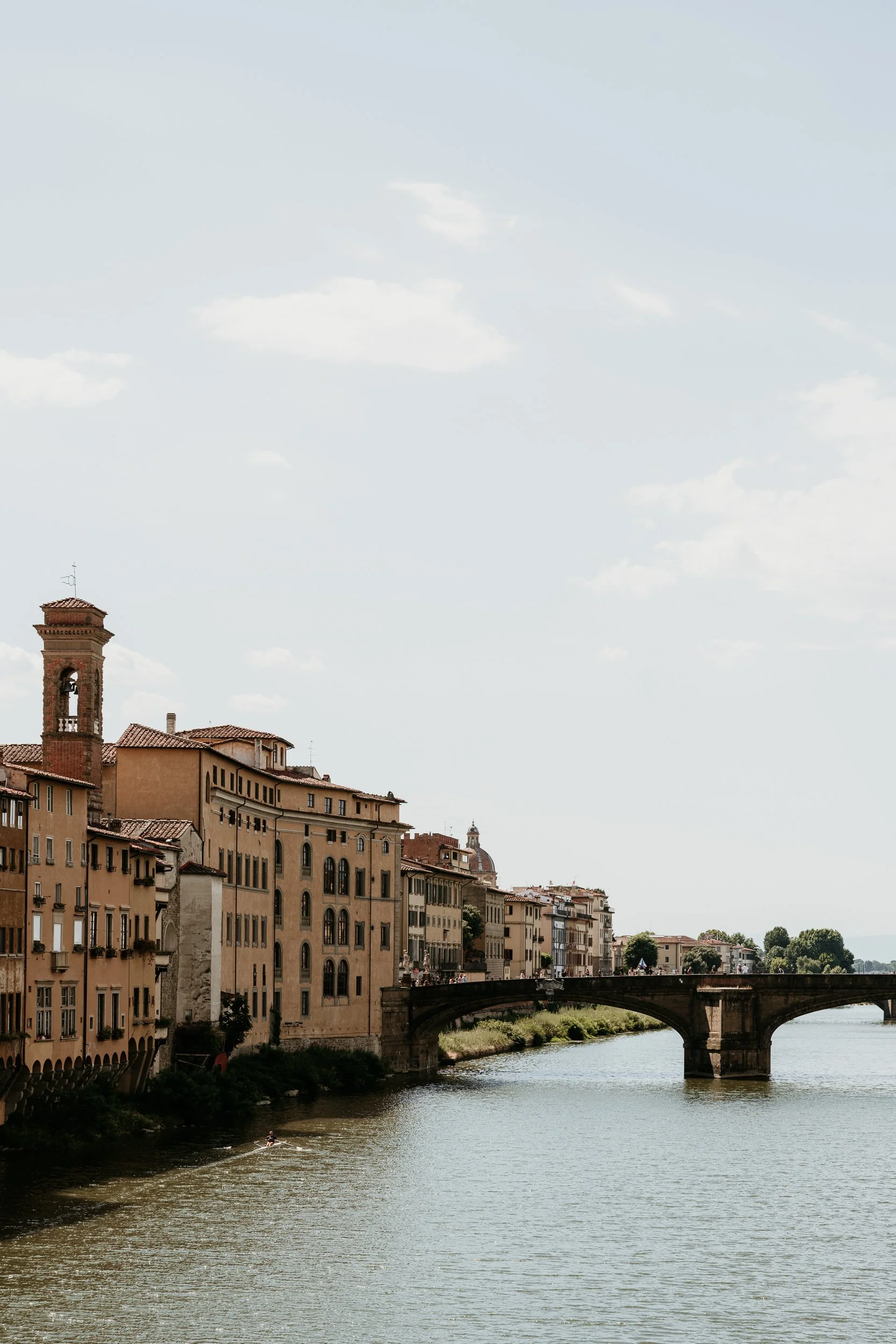 A scenic view of a river with a bridge and historic buildings along the riverside in a European city, under a partly cloudy sky.