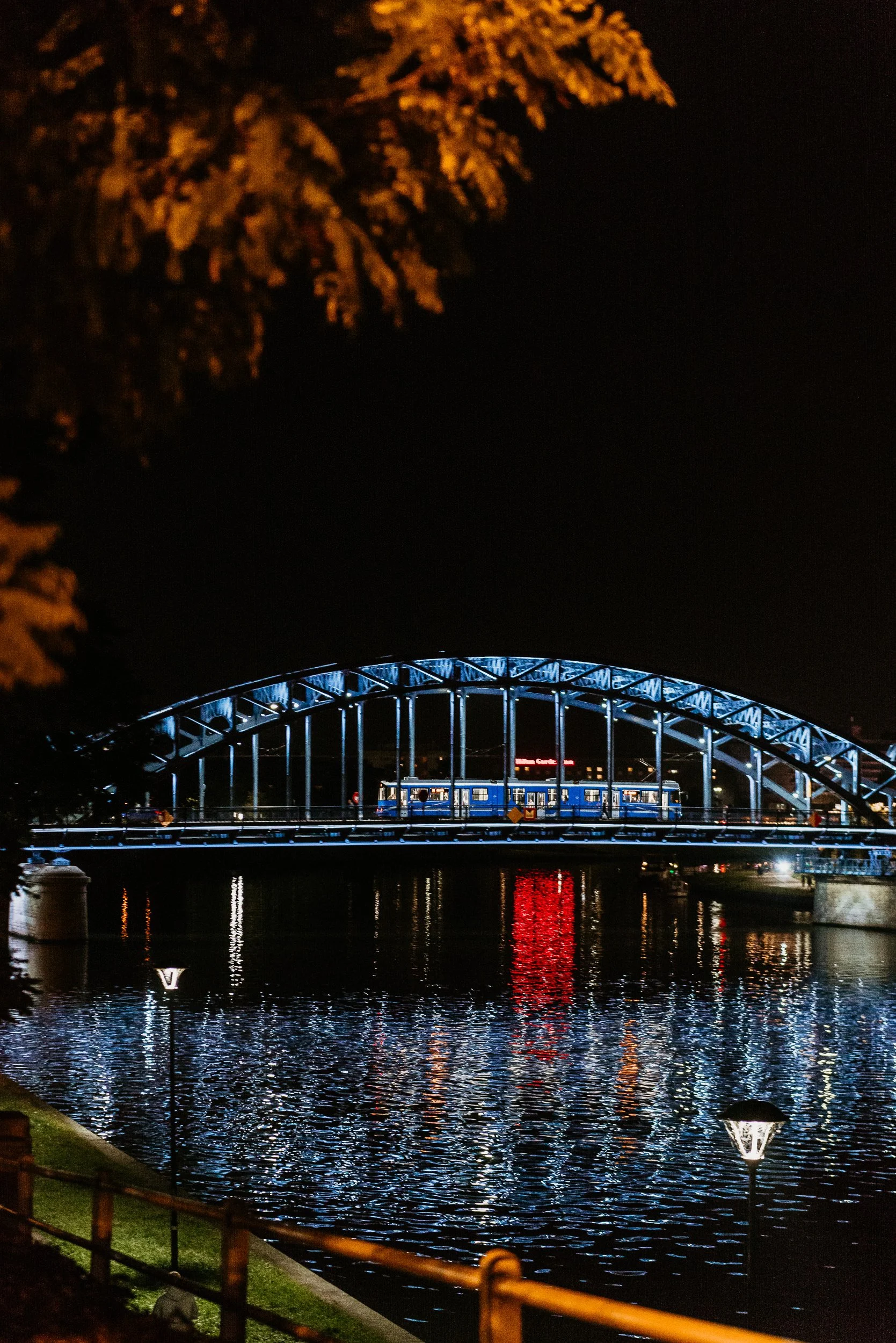 Nighttime view of a blue lit bridge over a river with reflections of the lights on the water. A train is crossing the bridge, and there are trees and streetlights along the riverbank.