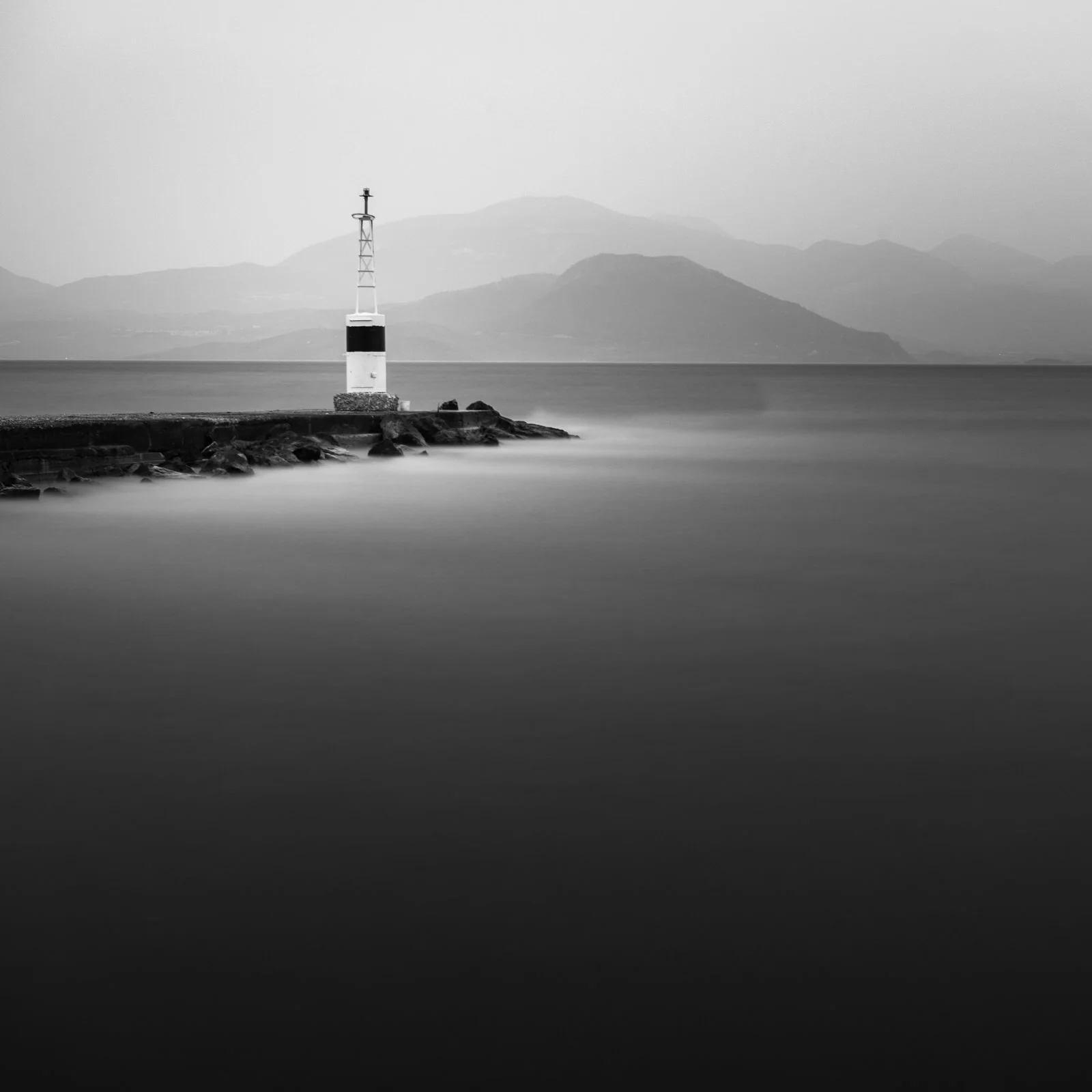 A black and white photo of a lighthouse on a rocky pier extending into calm water with mountains in the background.