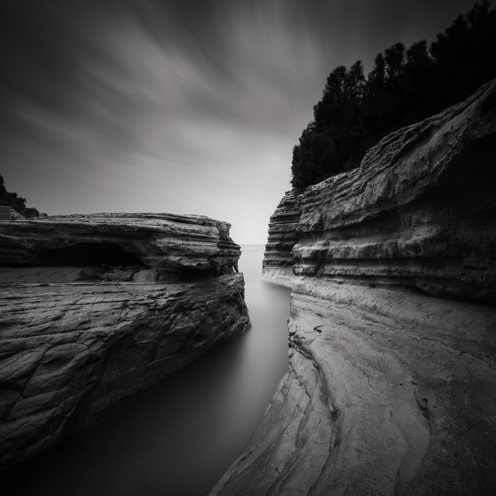 Black and white photo of a narrow water passage between layered rock formations with trees on a hillside in the background.