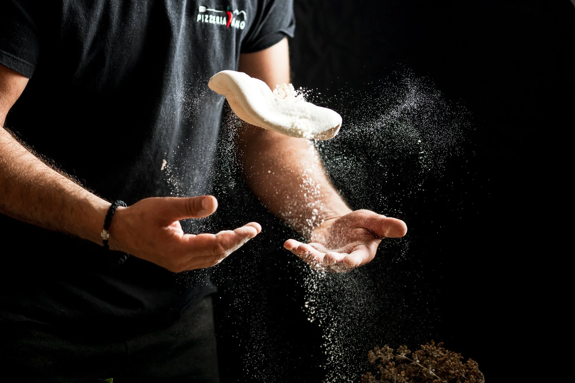 A person wearing a black T-shirt with a pizzeria logo is tossing pizza dough in the air, with flour in motion against a dark background. Hotels, Villas, Airbnb photography. Corfu, Greece.