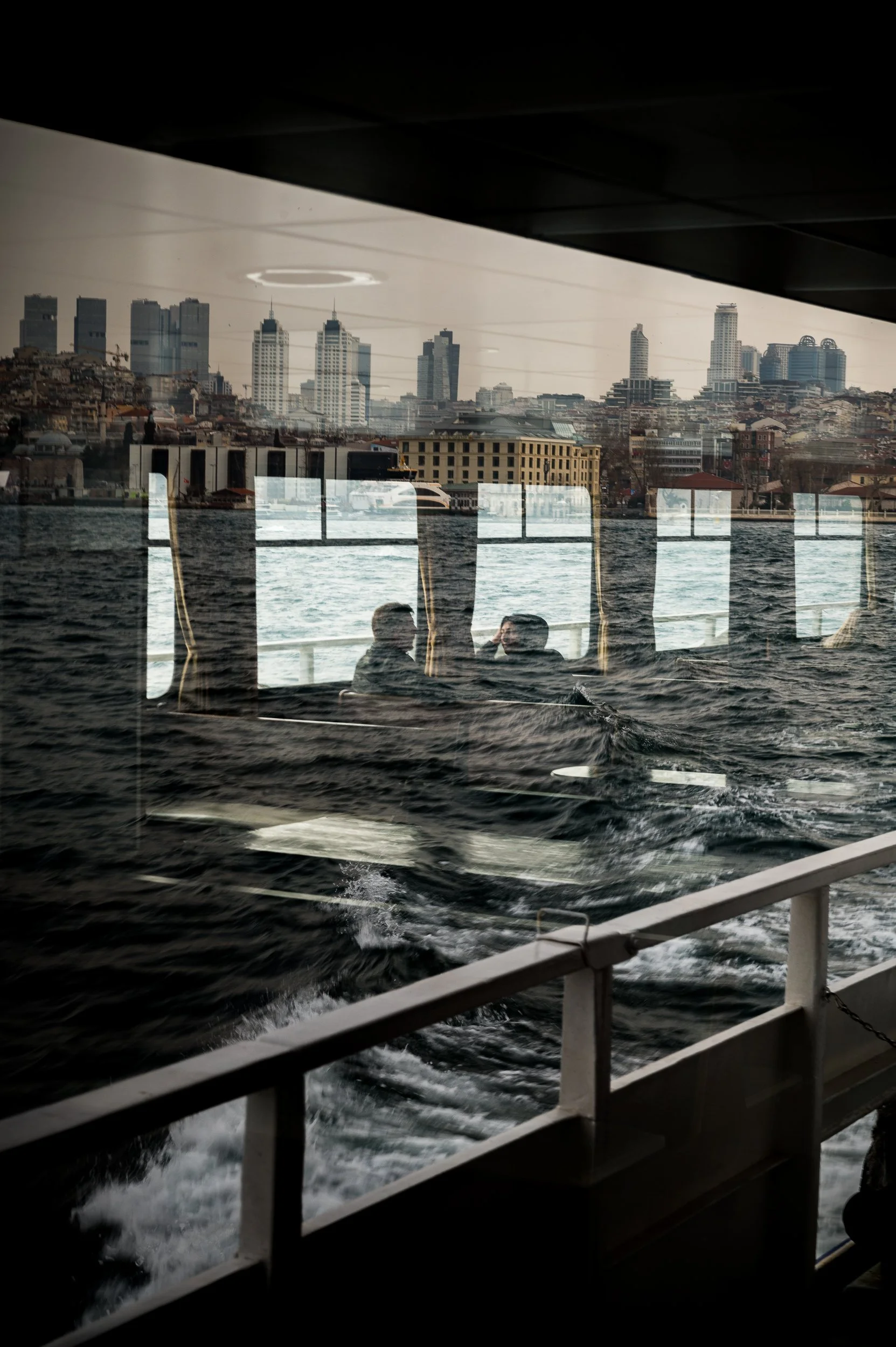 Two people sitting and talking on a boat with a city skyline and water visible through the window reflection.