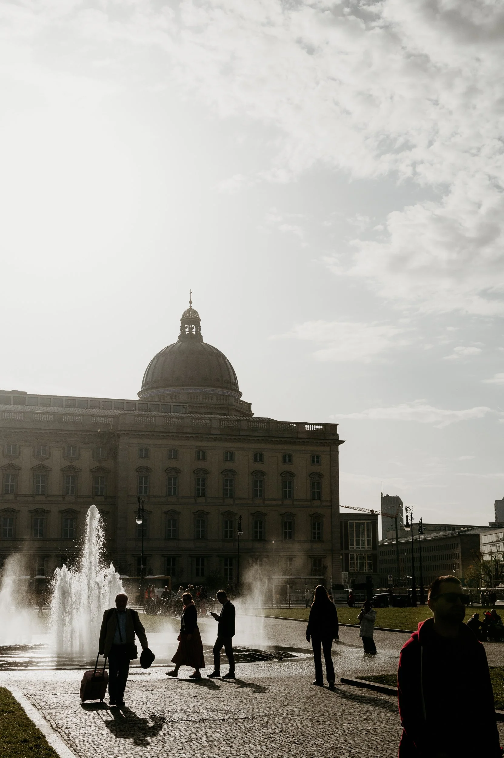 A city square with a fountain, people walking and standing, and a large historic building with a dome in the background, under a partly cloudy sky.
