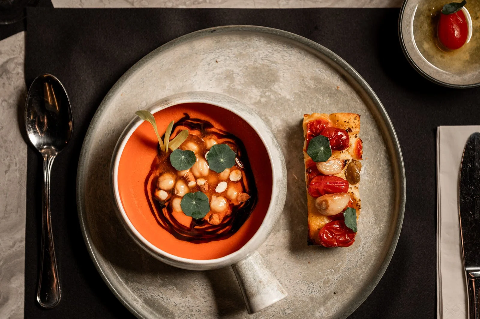 A tray holding a white bowl with an orange soup topped with white pearls, dark sauce, garnished with green leaves flowers, and a piece of bread with cherry tomatoes, olives, and herbs on top. Hotels, Villas, Airbnb photography. Corfu, Greece.