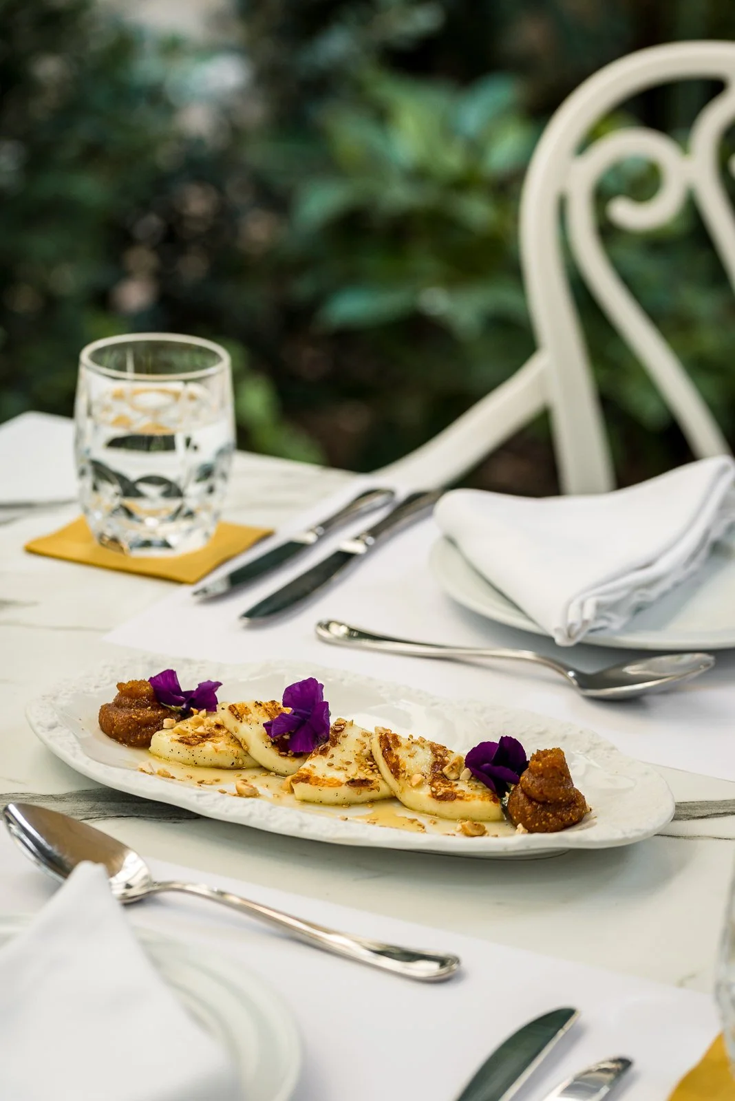Elegant table setting with a white plate of cheese garnished with purple edible flowers and nuts, a glass of water on a coaster, silverware, a white napkin, and a blurred garden background. Hotels, Villas, Airbnb photography. Corfu, Greece.