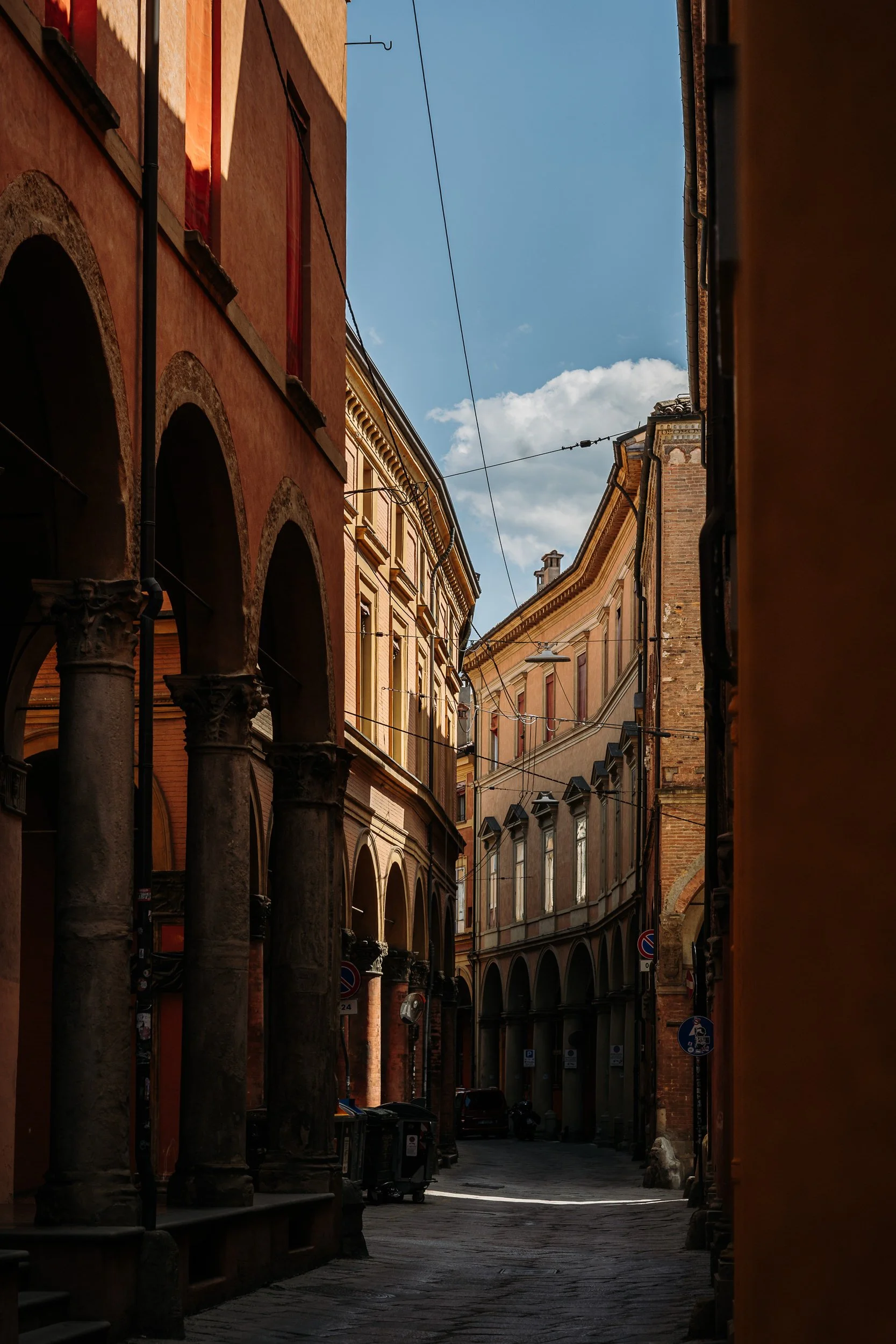 A narrow European street with historic buildings, arches on the left, and overhead wires, under a blue sky with clouds.