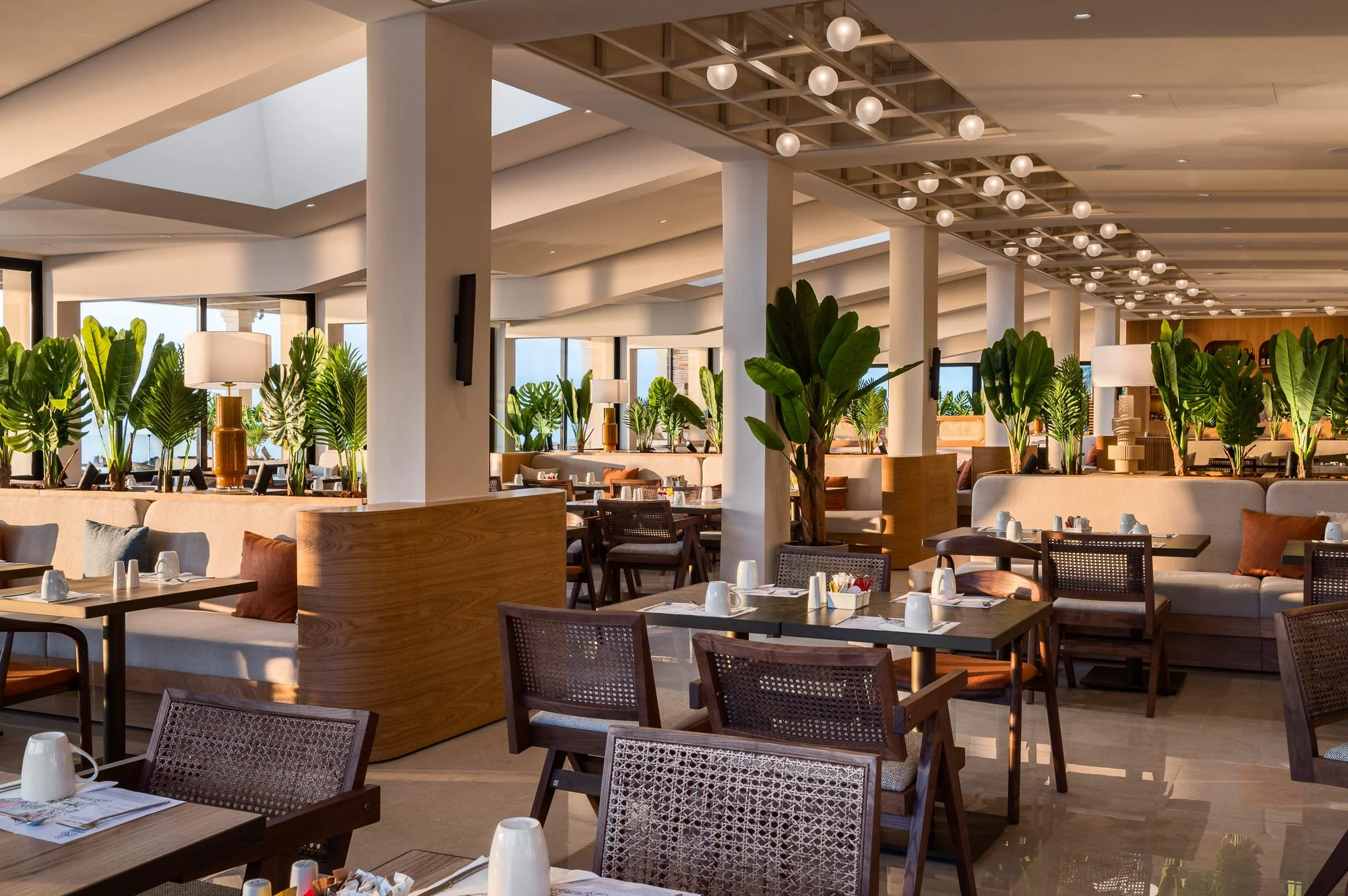 Elegant restaurant with wooden furniture, large green potted plants, table settings, and modern lighting, bright with natural sunlight. Hotels, Villas, Airbnb photography. Corfu, Greece.