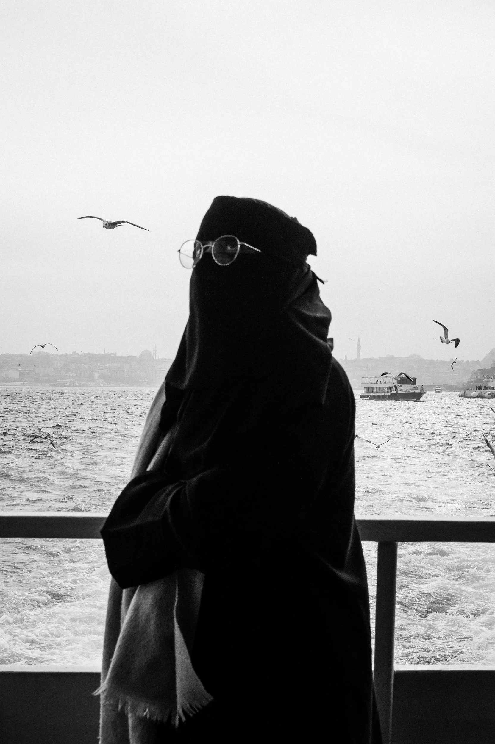A person dressed in black with a head covering and glasses standing by a water railing, with seagulls flying around and boats in the background on a waterfront.
