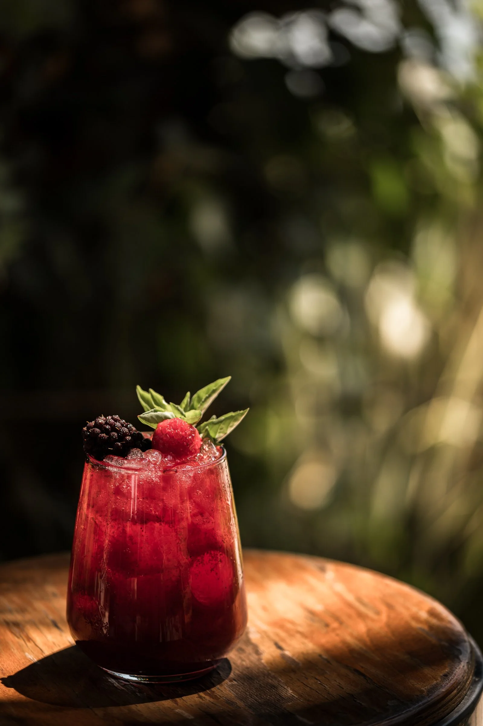 A glass of berry cocktail garnished with fresh mint leaves and mixed berries, sitting on a wooden surface with sunlight and outdoor blurred greenery in the background. Hotels, Villas, Airbnb photography. Corfu, Greece.
