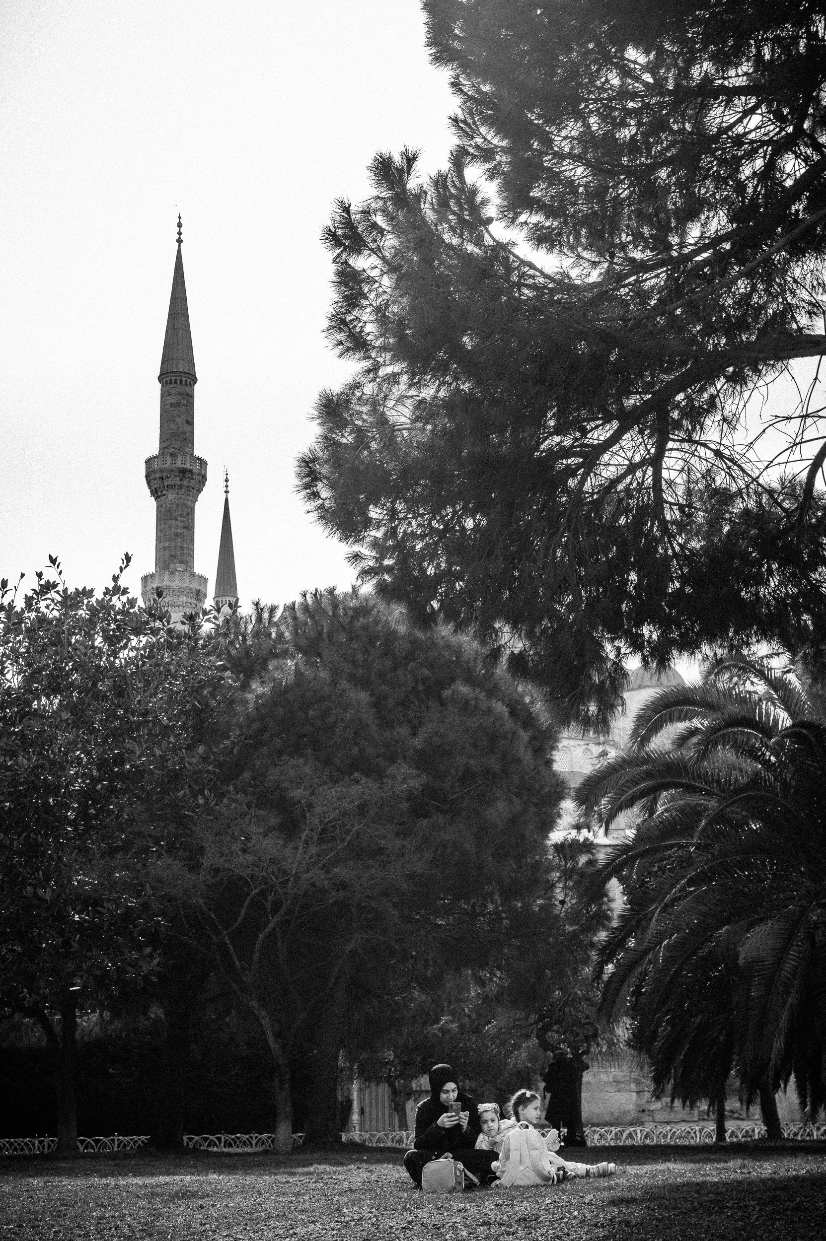 A woman and two children sitting on the grass in a park, surrounded by trees with a tall minaret and a mosque in the background, in black and white.