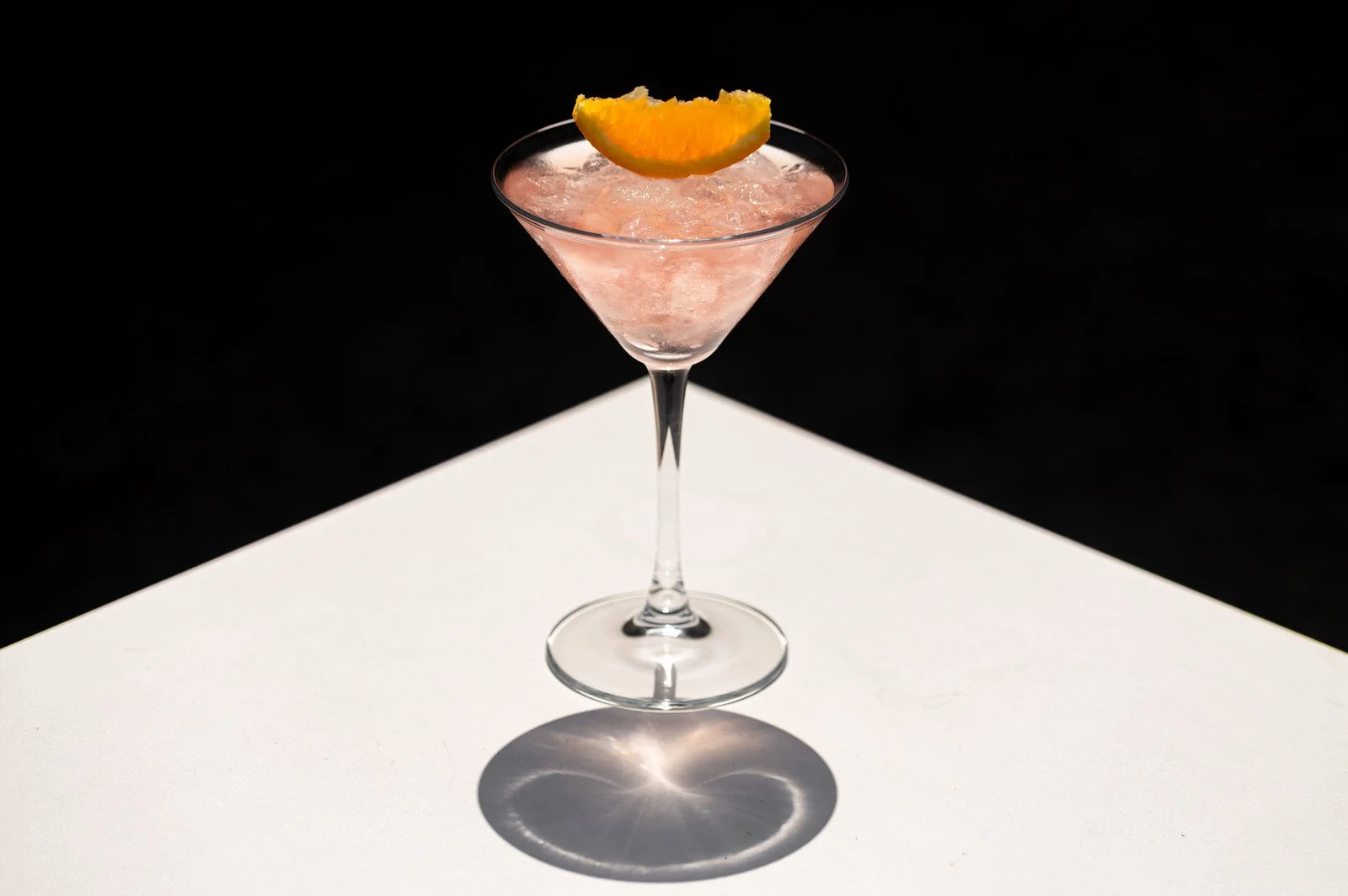 A pink cocktail with crushed ice in a martini glass, garnished with a slice of orange on the rim, on a white surface with a shadow and reflection.Hotels, Villas, Airbnb photography. Corfu, Greece.