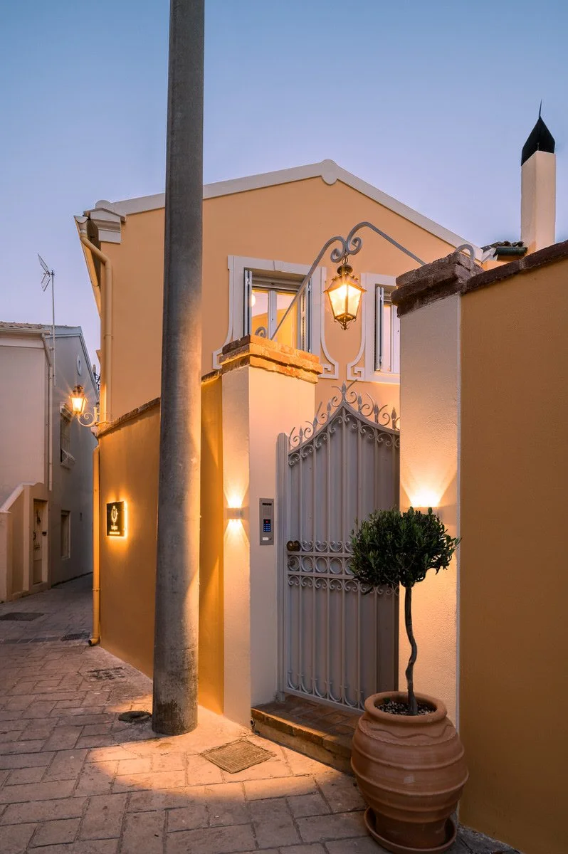 A charming house with a yellow exterior and white trim, lit by warm outdoor lamps, has a decorative white metal gate and a potted plant next to it. With clear evening sky overhead. Hotels, Villas, Airbnb photography. Corfu, Greece.
