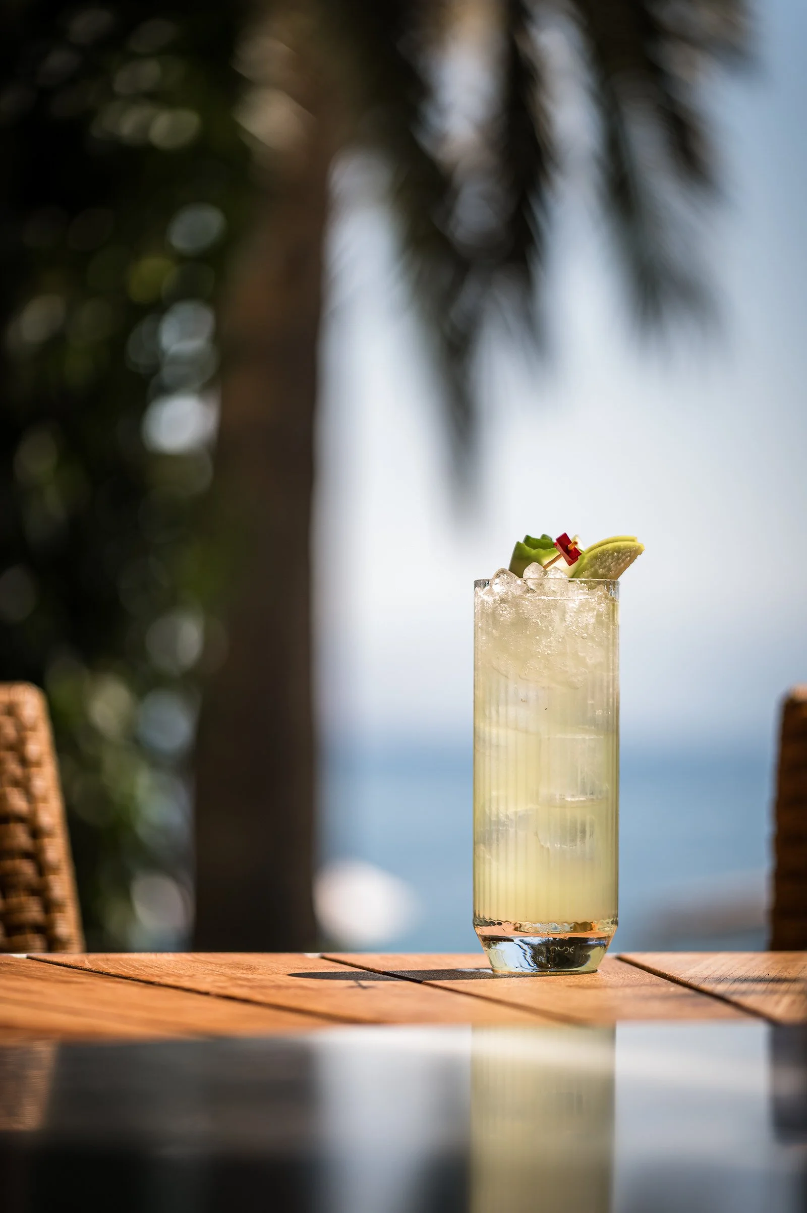 A tall glass of lemonade with ice, garnished with lime wedge, cherry, and green fruit, sitting on a wooden table outdoors with blurred trees and ocean in the background.Hotels, Villas, Airbnb photography. Corfu, Greece.