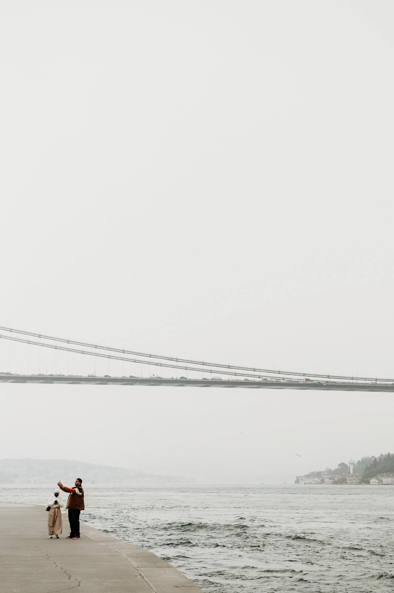 Two people standing on a concrete pathway by the water, with a large suspension bridge in the background and a cloudy sky.