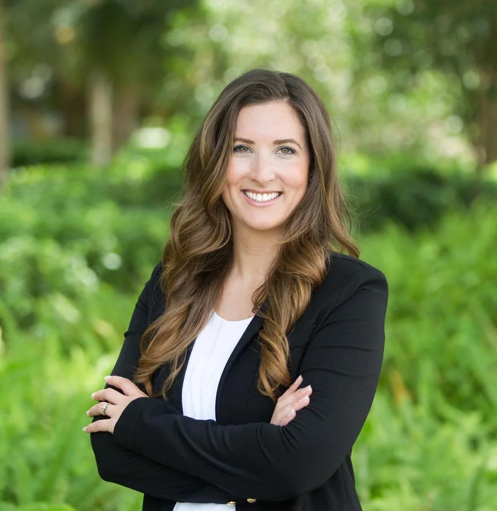 A woman with long wavy brown hair, smiling and crossing her arms, stands outdoors in a lush green background with trees and foliage.