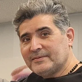 Close-up van een man met grijs haar en een glimlach, tegen een muur.